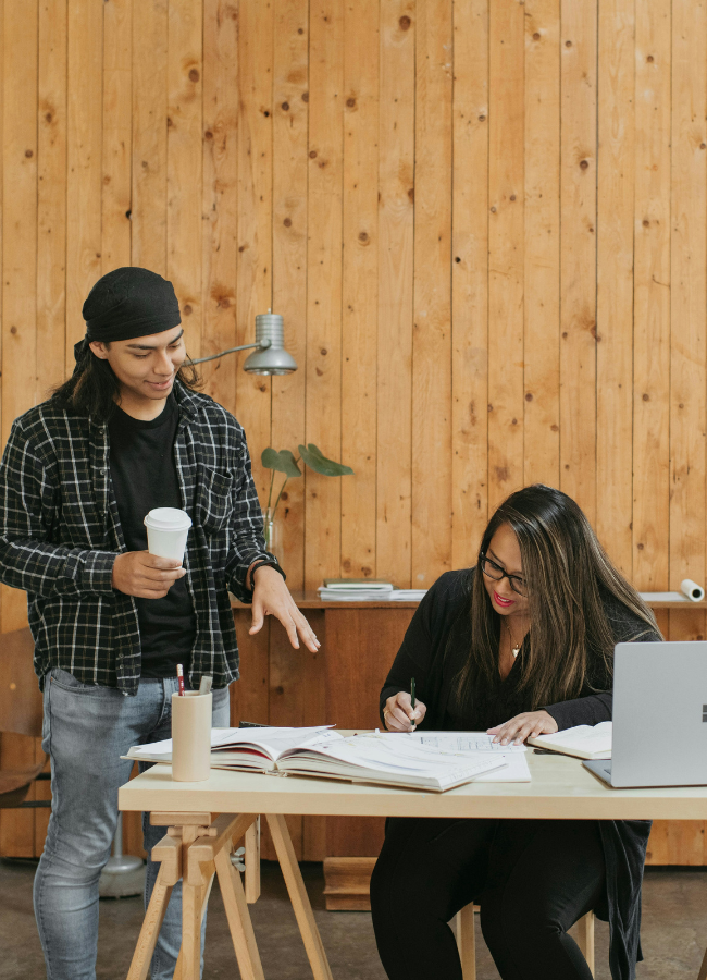 Photo of man and woman having a conversation next to a planner. He is standing, holding a coffee, and speaking, she is making notes.