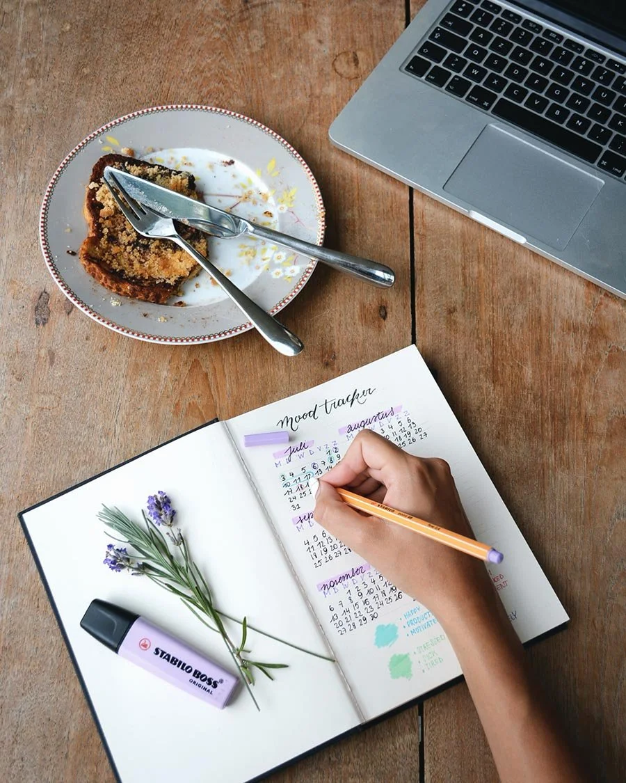 Photo of person writing in their mood journal, with cake and a laptop on the table