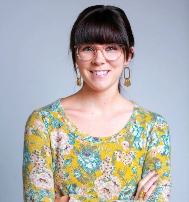 A woman with short dark hair and glasses smiling, wearing a floral patterned yellow dress and gold earrings.