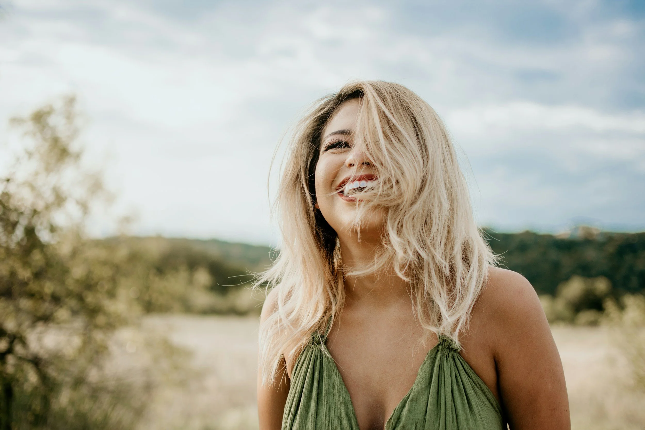 Young adult woman smiling with teeth showing, red lipstick, blond shoulder length hair with dark roots and wearing a green tank top.