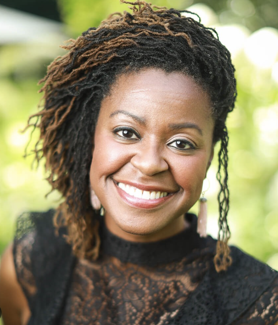 A smiling BIPOC woman in an outdoor setting with blurred green foliage background.