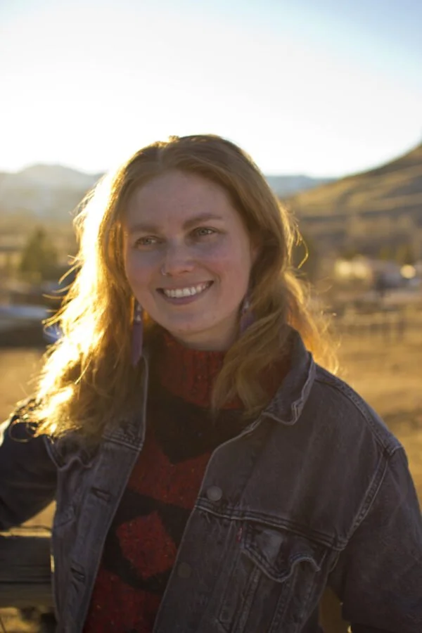 A woman with red hair smiling outdoors during sunset in a rural landscape with hills and scattered buildings.