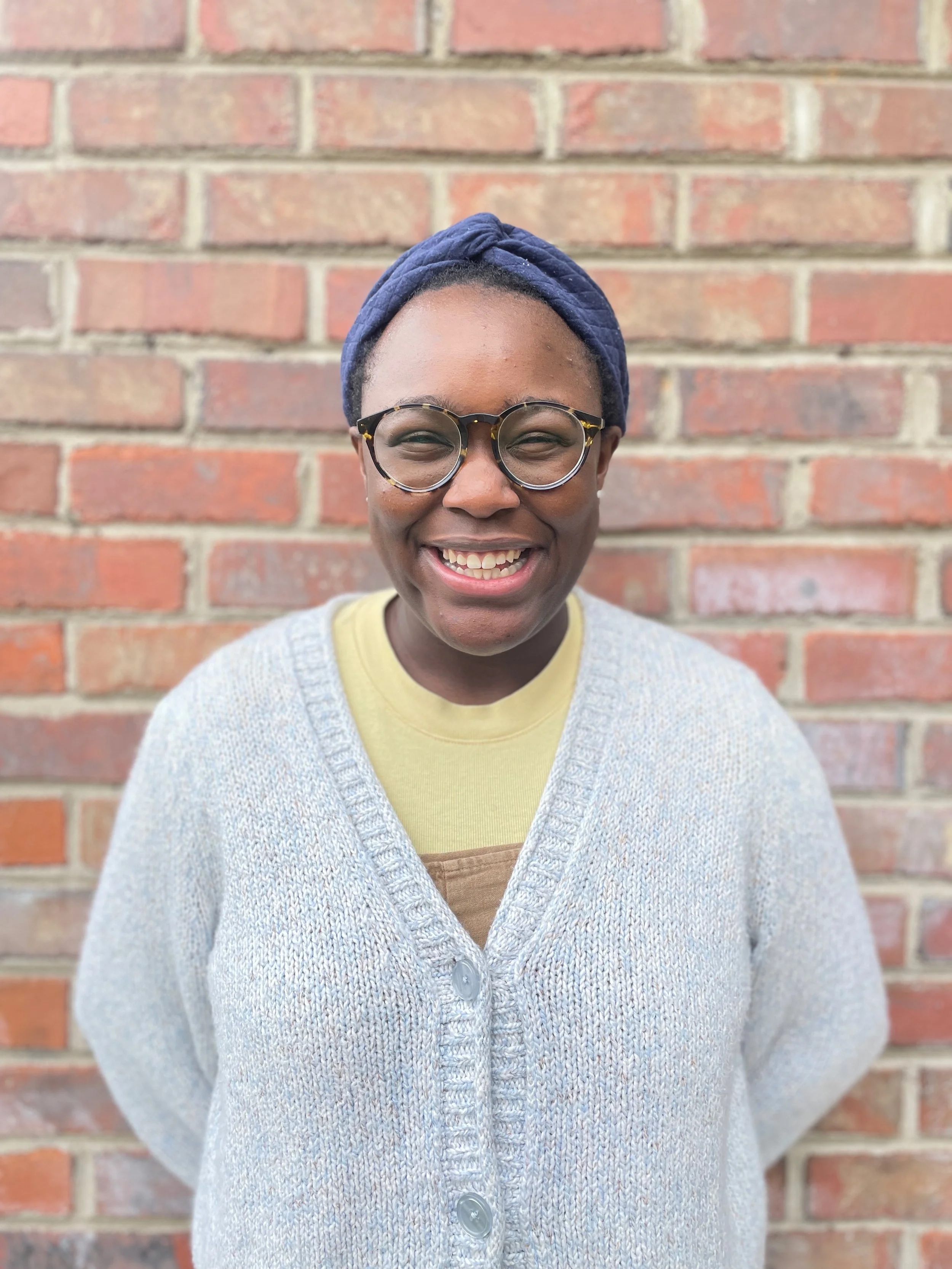 Image of OCD Therapist Reece Thomas against a brick wall. Reece is wearing glasses, a headscarf and grey cardigan.