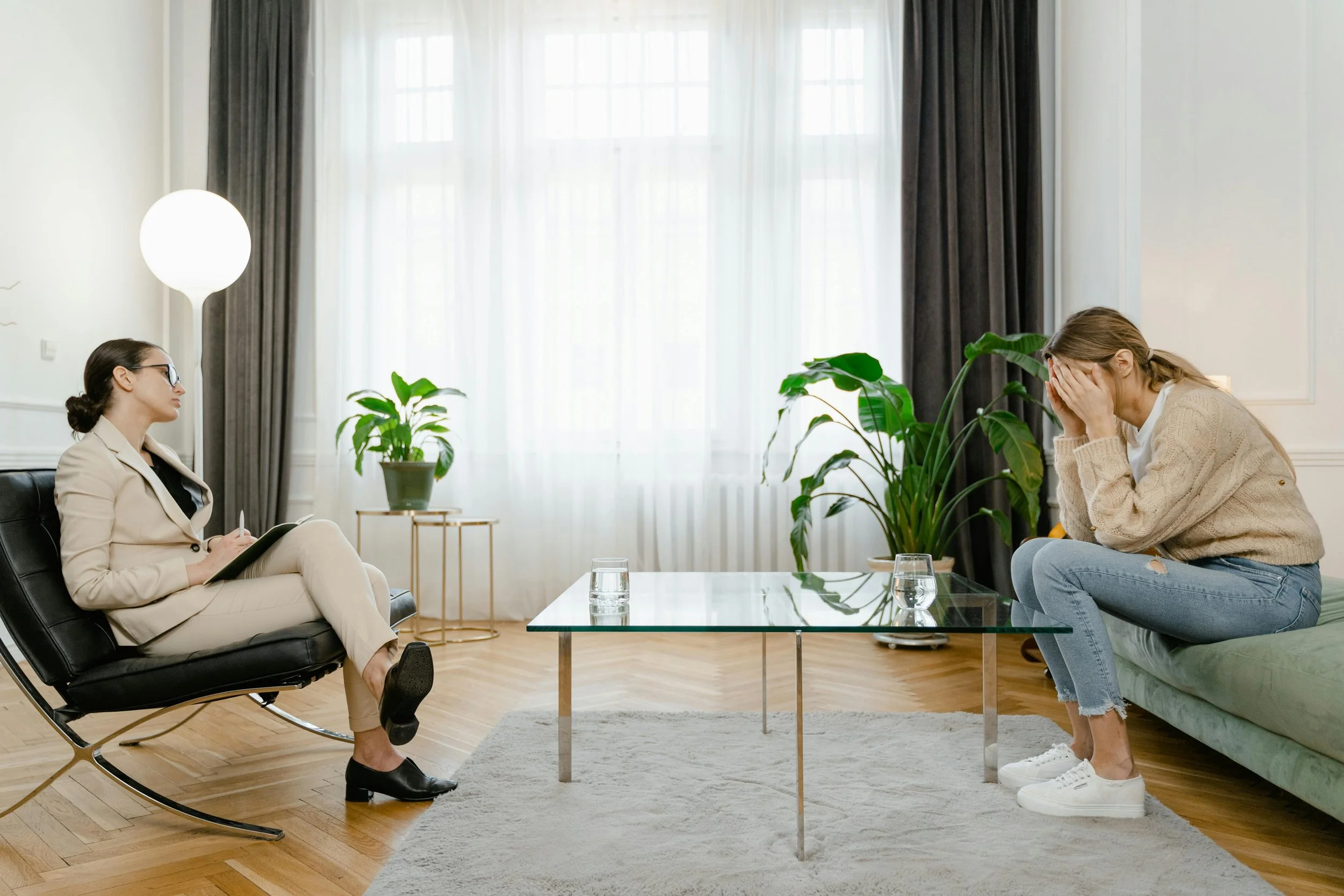 Female presenting therapist with female presenting client, sitting across from each other in an office setting, plants and natural light in the background, glass coffee table in between them.
