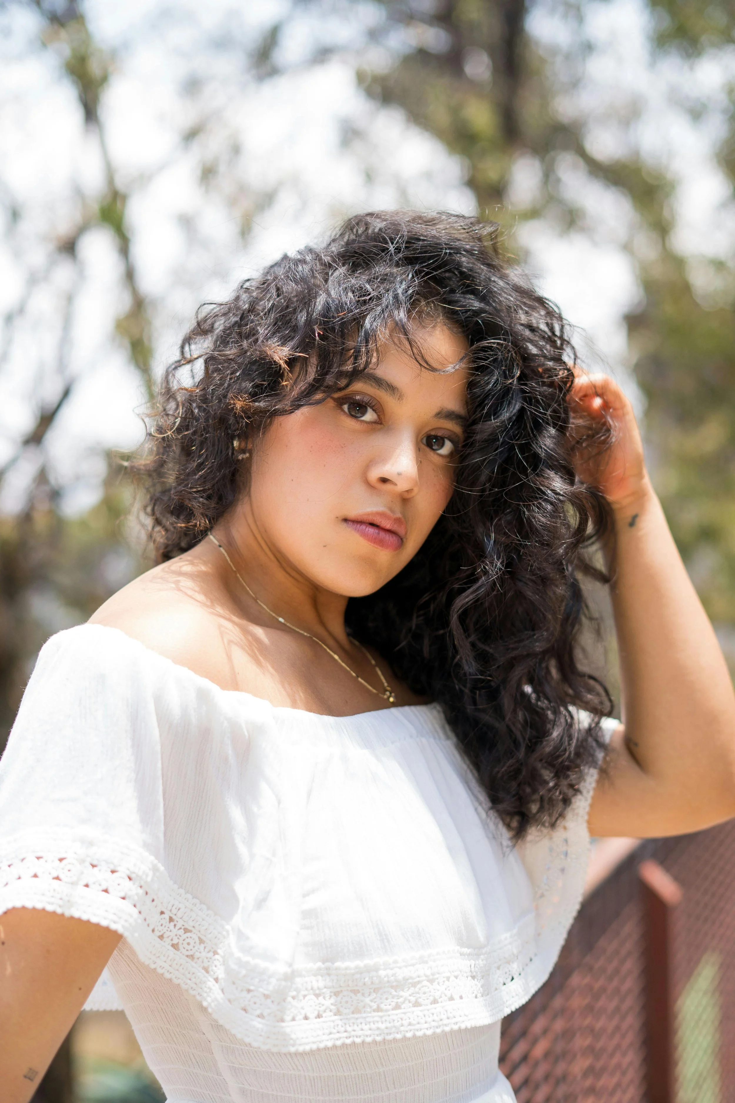 Latina woman with dark curly hair, wearing a white off the shoulder top with a gold necklace, tree in the background with natural lighting.