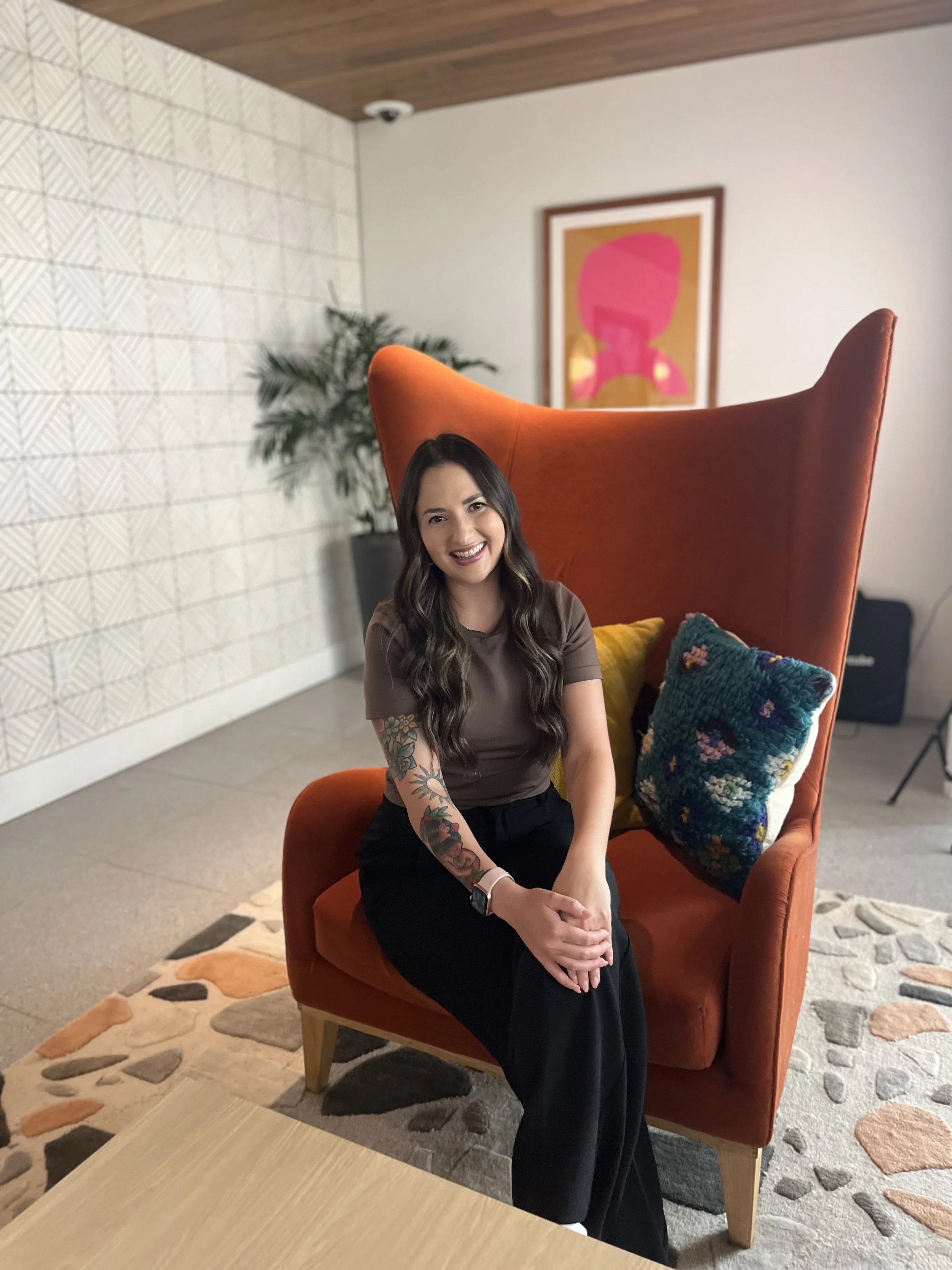 Eating disorder therapist Allyson Ford sitting on an orange chair with teal patterned pillows, abstract art and a plant in the background