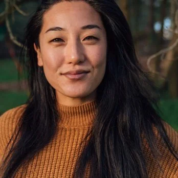 Close-up of a woman with long black hair, wearing an orange-brown sweater, outdoors with blurred trees in the background.