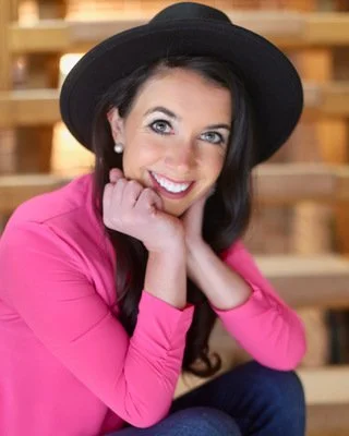 A woman wearing a black hat and pink top, smiling with her chin resting on her hand, sitting against a wooden background.