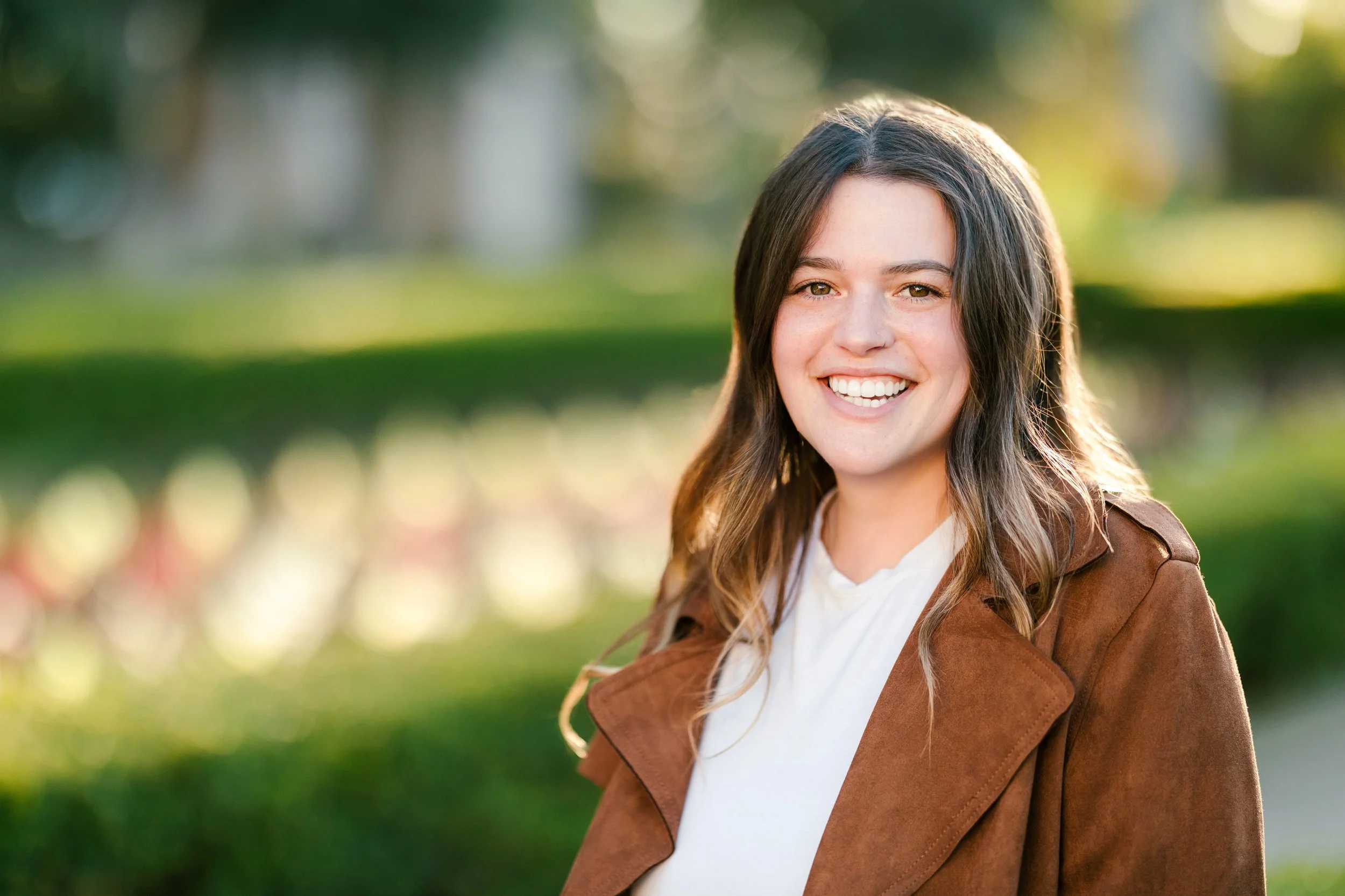 Georgia, an eating disorder therapist, sits in Balboa Park in a brown jacket and is smiling