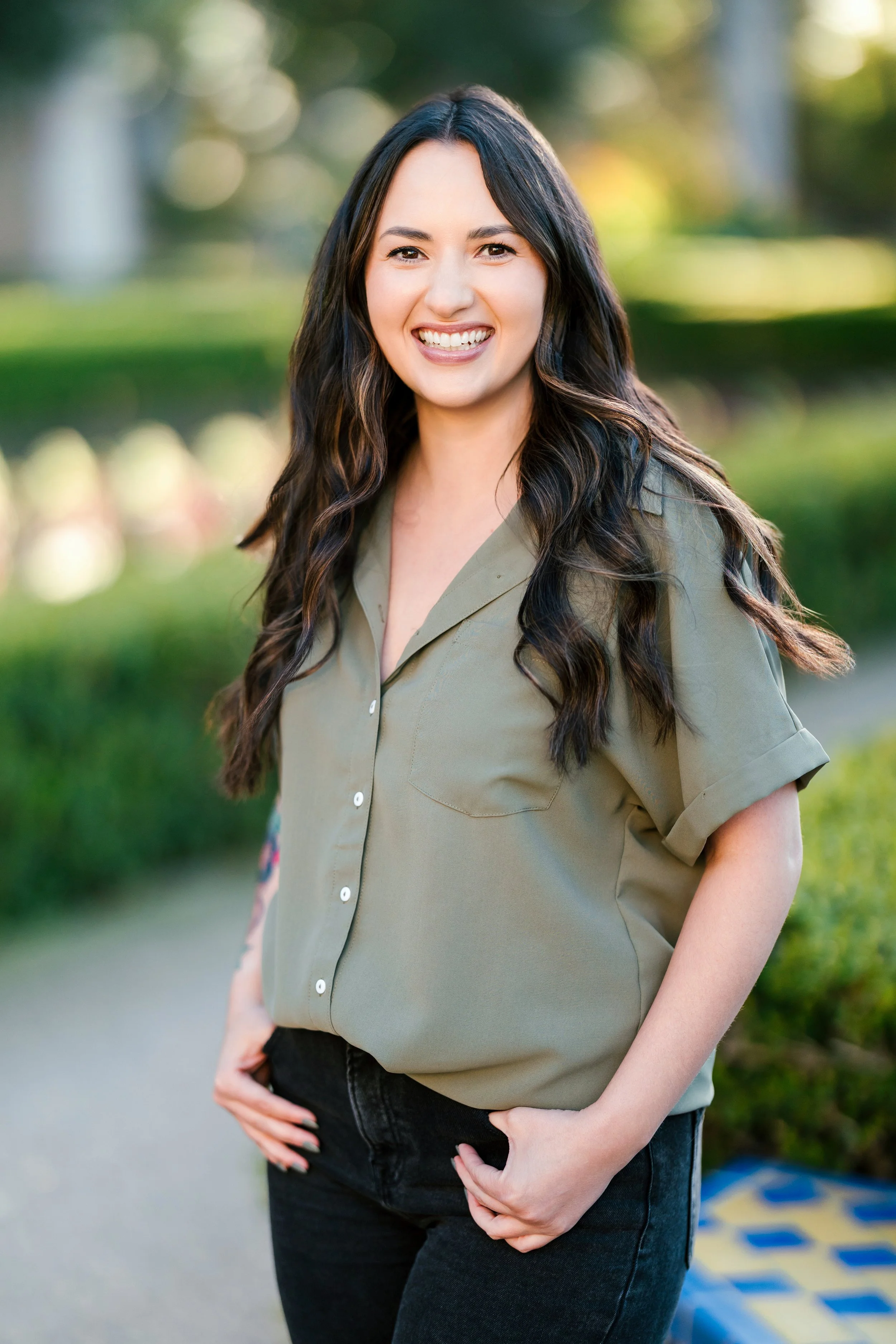 A woman smiling outdoors with a blurred green background, wearing a khaki shirt and black pants.