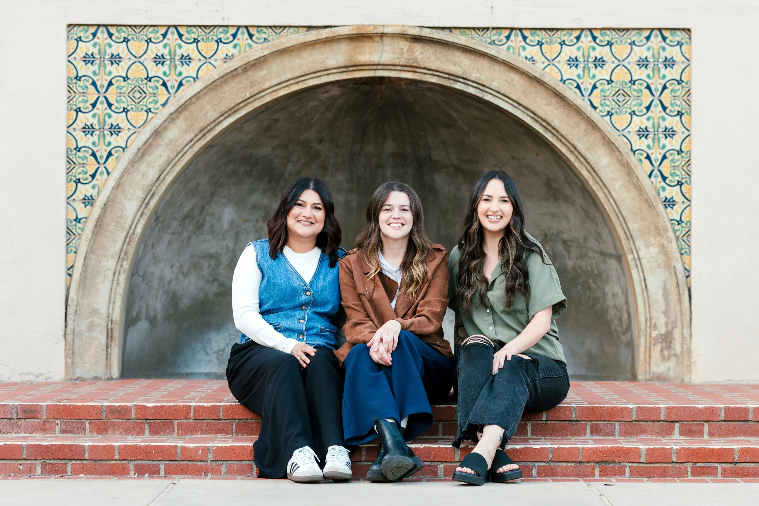 Three young women sitting on red brick steps in front of an arched alcove with colorful tile decorations, smiling at the camera.