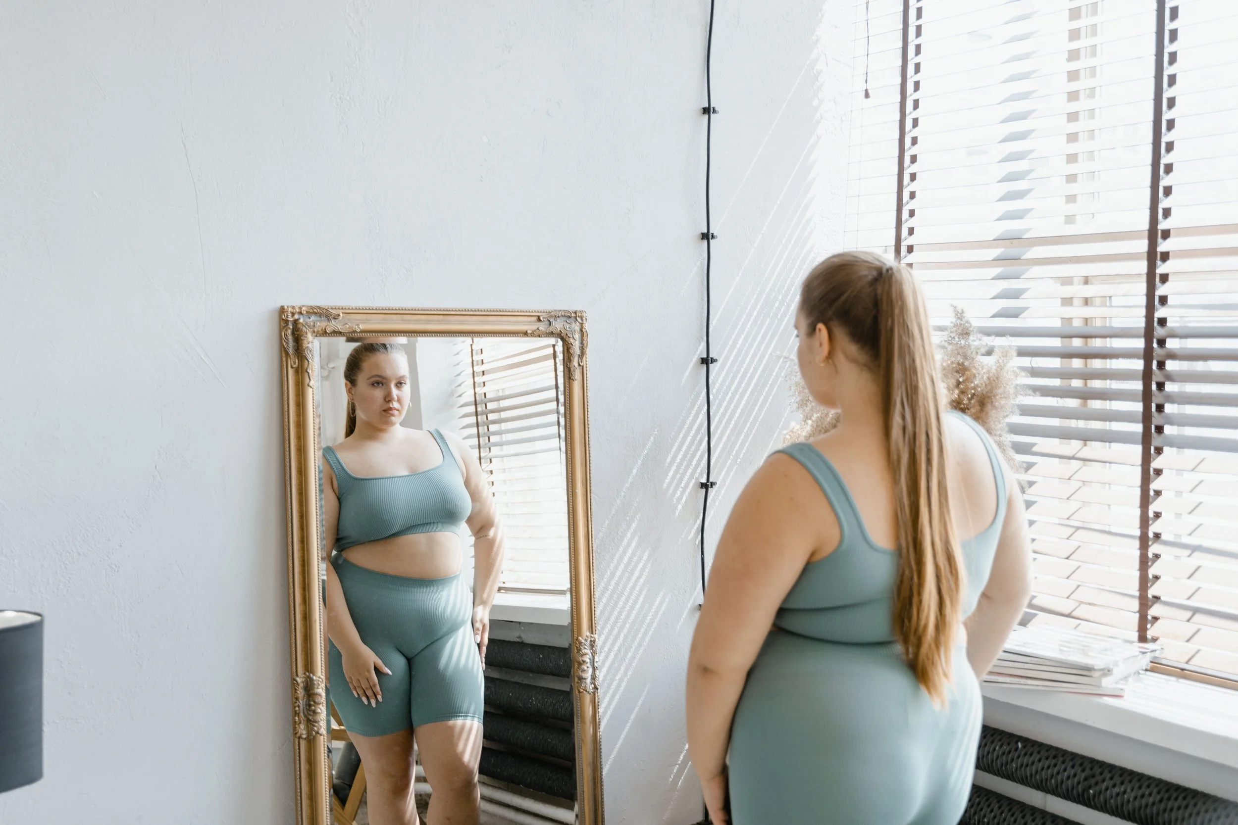 Mid size, Caucasion young woman looking at herself in the mirror in a matching, light blue set.