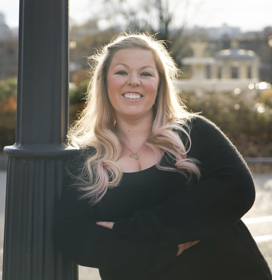 A smiling woman with long wavy blonde hair standing outdoors near a lamp post, with trees and buildings in the background during late afternoon sunlight.