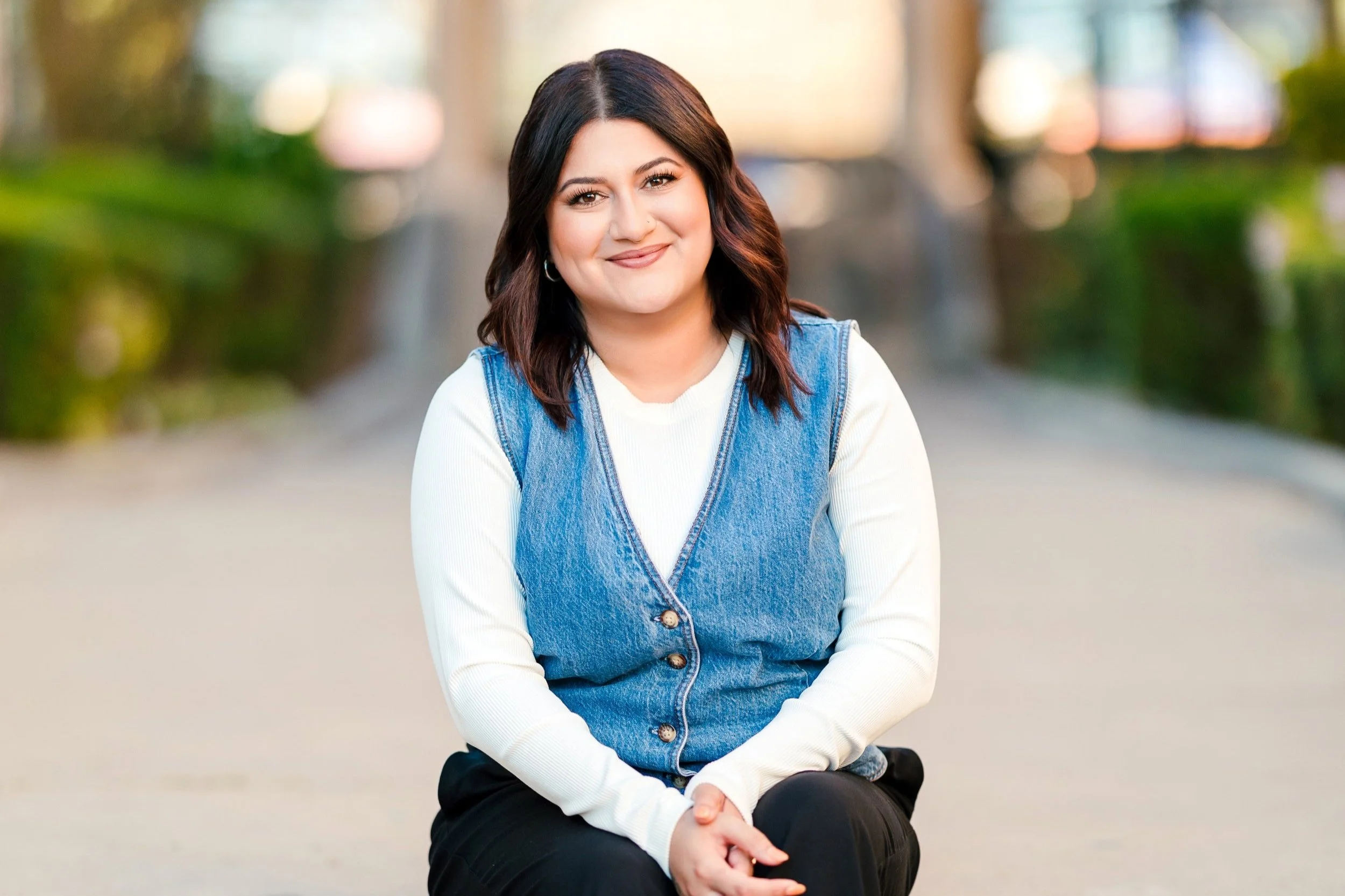 OCD therapist with shoulder-length dark brown hair sitting outdoors on a paved surface. She is wearing a white long-sleeve shirt underneath a sleeveless denim vest, and black pants. The background is blurred with greenery and trees.