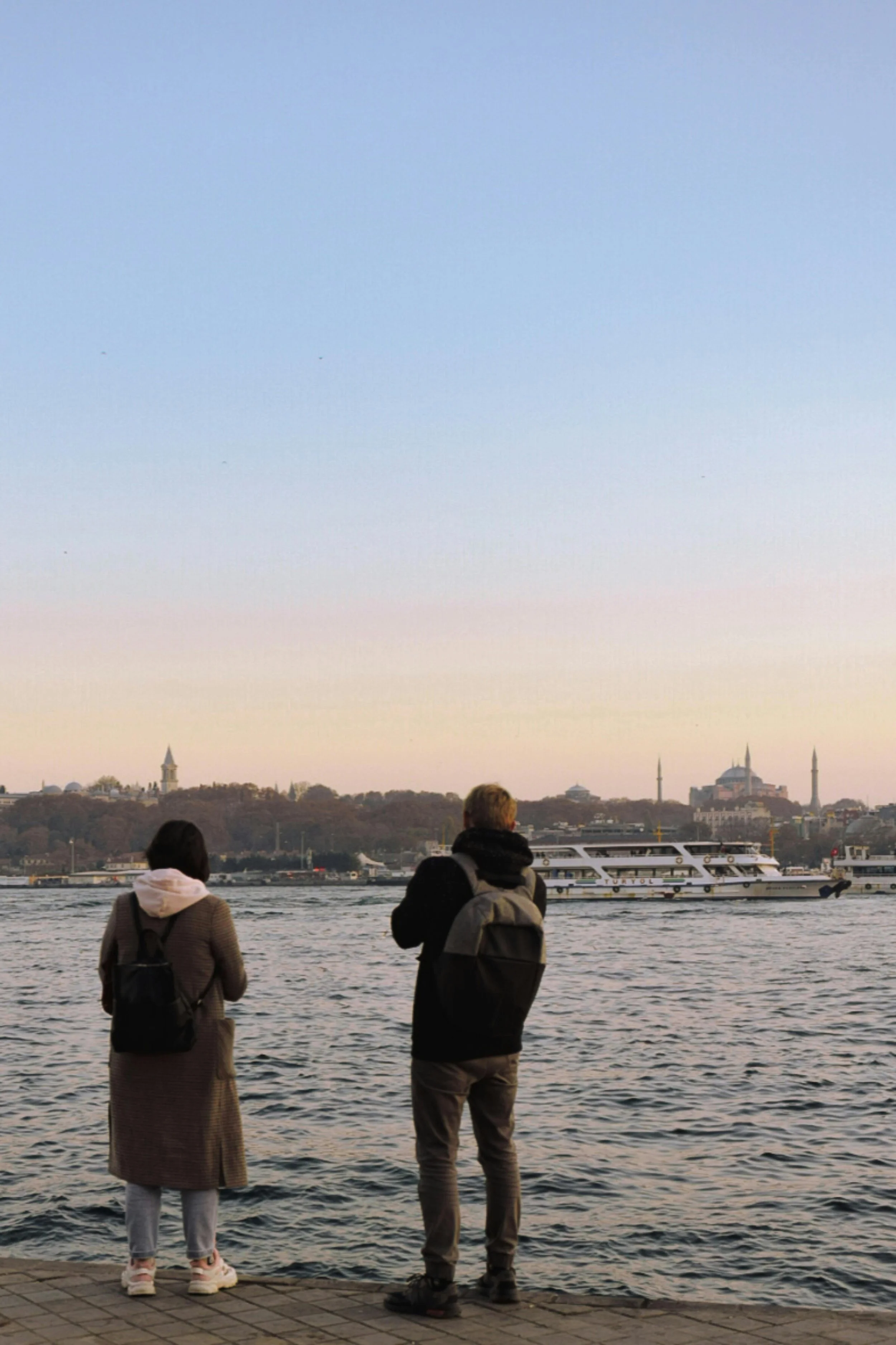 two people with backpacks, looking towards a body of water. Appearing to be traveling.