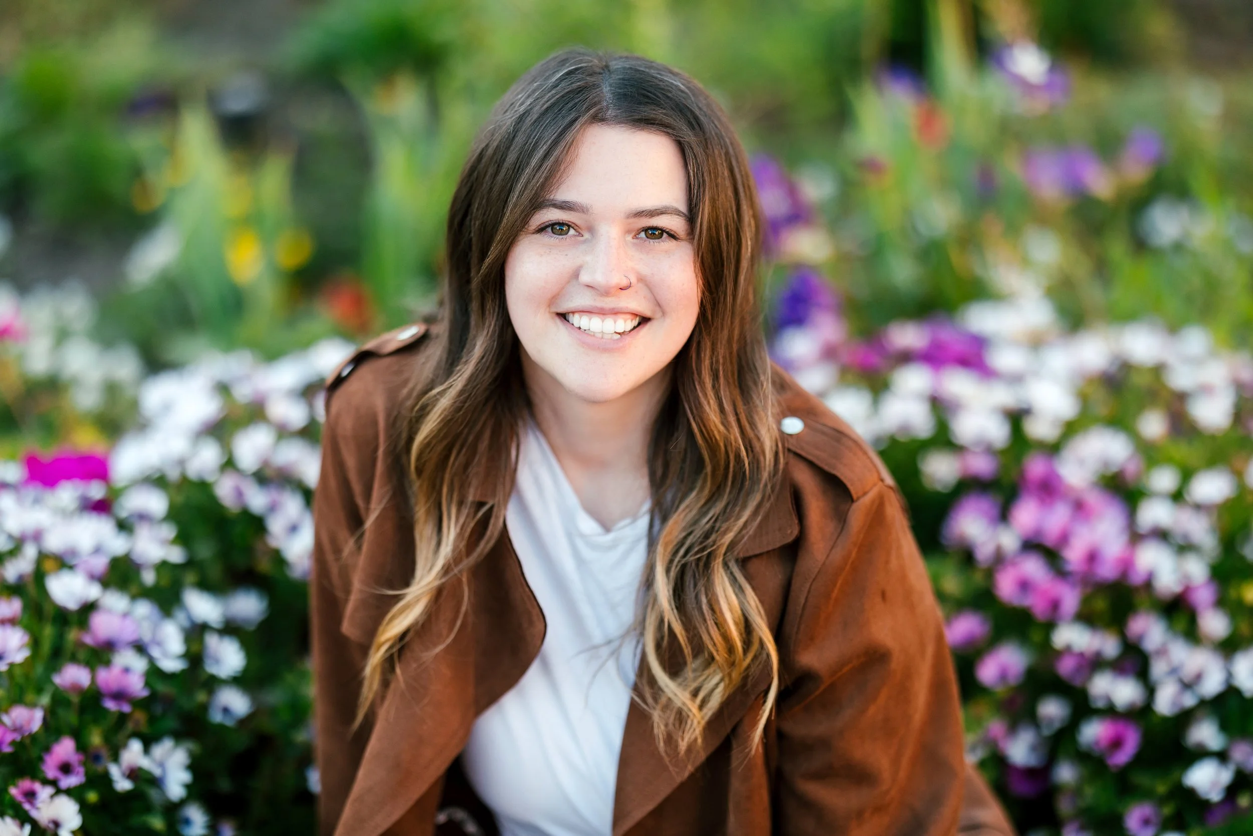 Eating Disorder therapist with long wavy brown hair smiling outdoors among pink, purple, and white flowers, wearing a brown jacket over a white top.