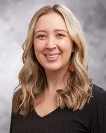 A woman with long wavy blonde hair smiling at the camera, wearing a black top and orange earrings, against a gray background.