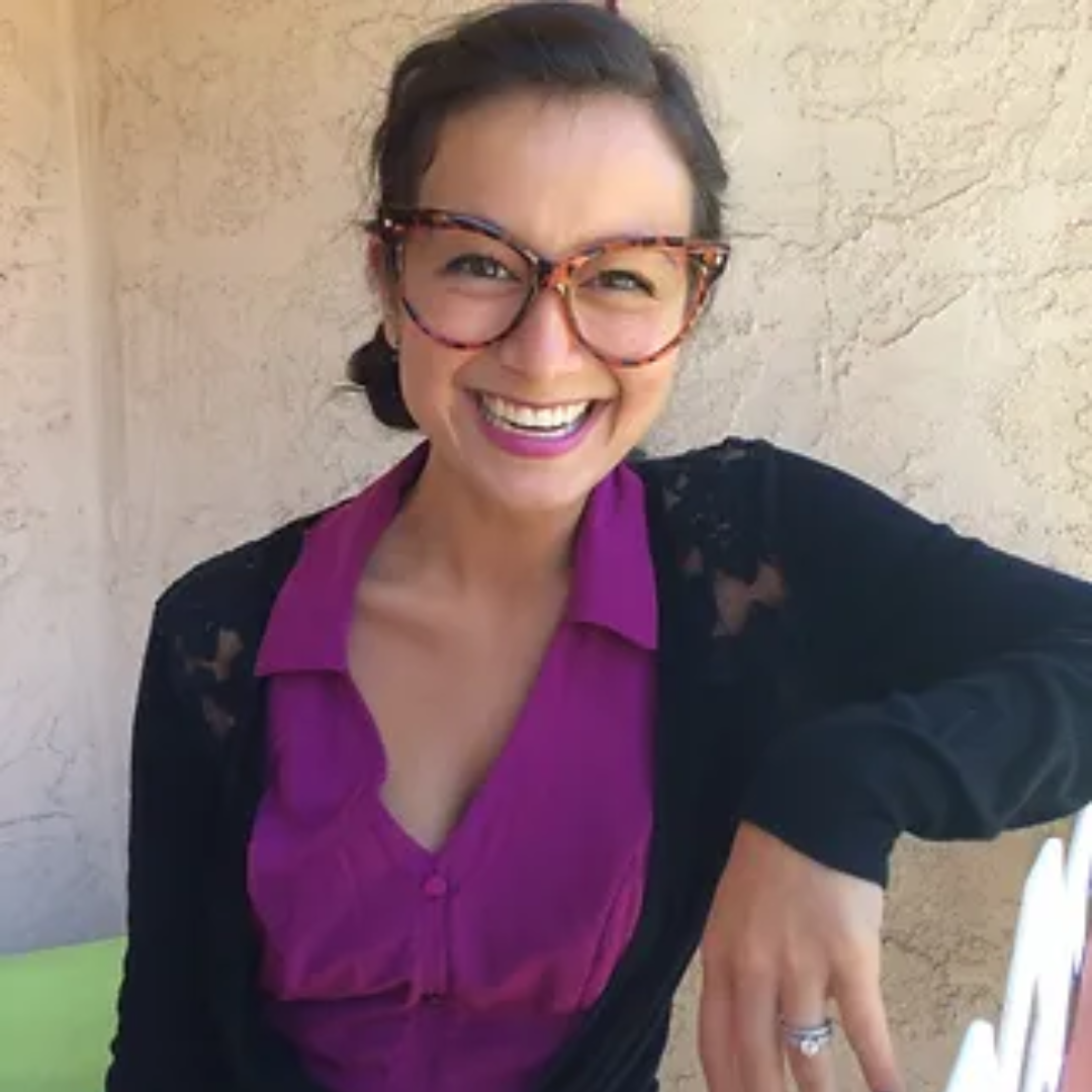 A woman with short dark hair, wearing glasses, a purple blouse, and a black sweater, smiling and showing her teeth, seated against a beige textured wall.