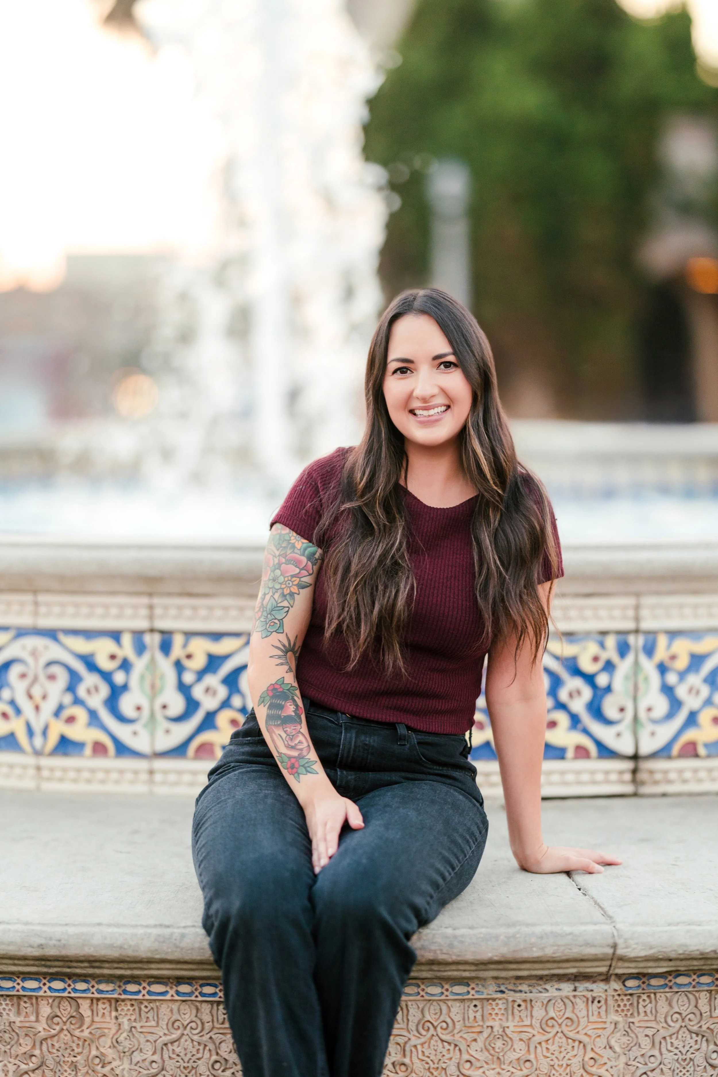 A female adult eating disorder and OCD therapist with long dark hair and tattoos on her left arm, wearing a maroon shirt and black pants, sitting on the edge of a decorative fountain with water in the background, outdoors during daytime, in San Diego
