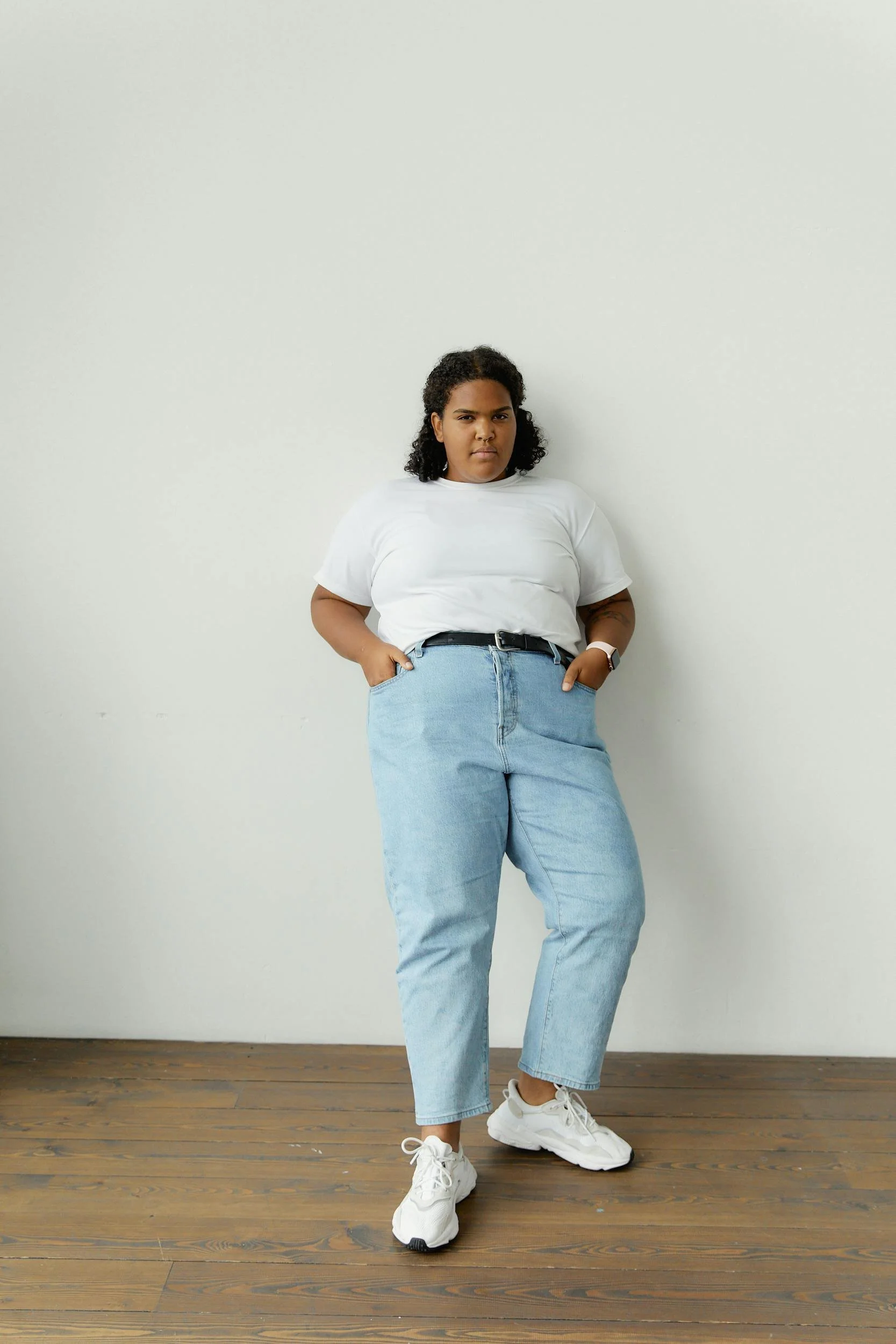 Woman of color with dark curly, shoulder length hair, standing against a white wall and wearing a white shirt, dark belt and light jeans with white sneakers on hardwood floor. She has her hands in her pocket and appears to be in a serious mood.