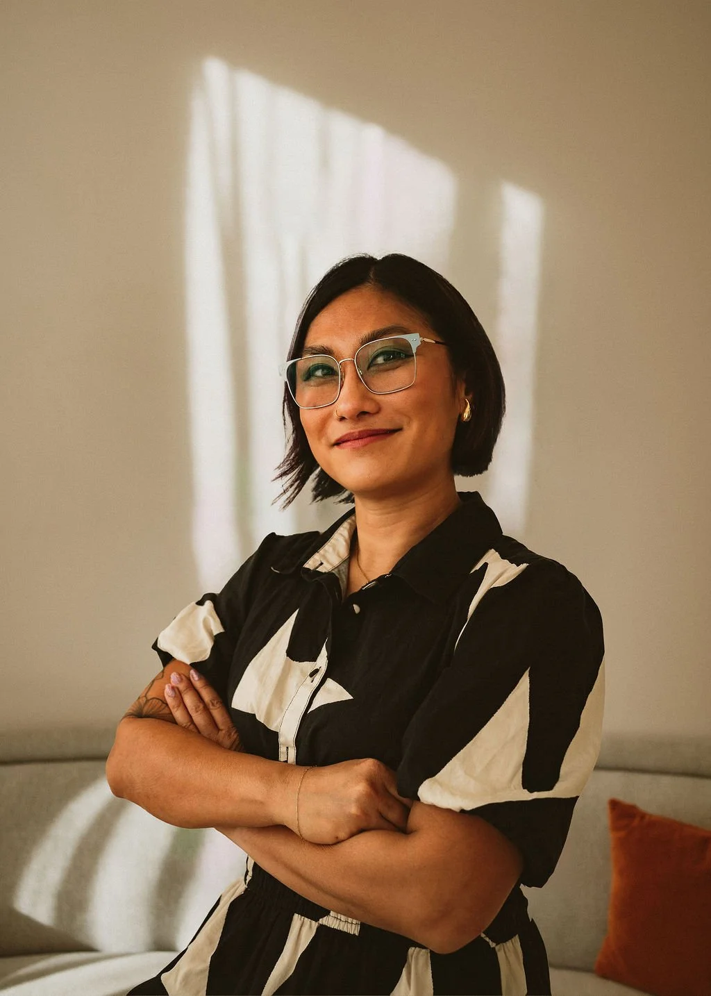 A woman with short black hair, glasses, and earrings, standing with arms crossed in front of a wall with a shadow of a window and sunlight.