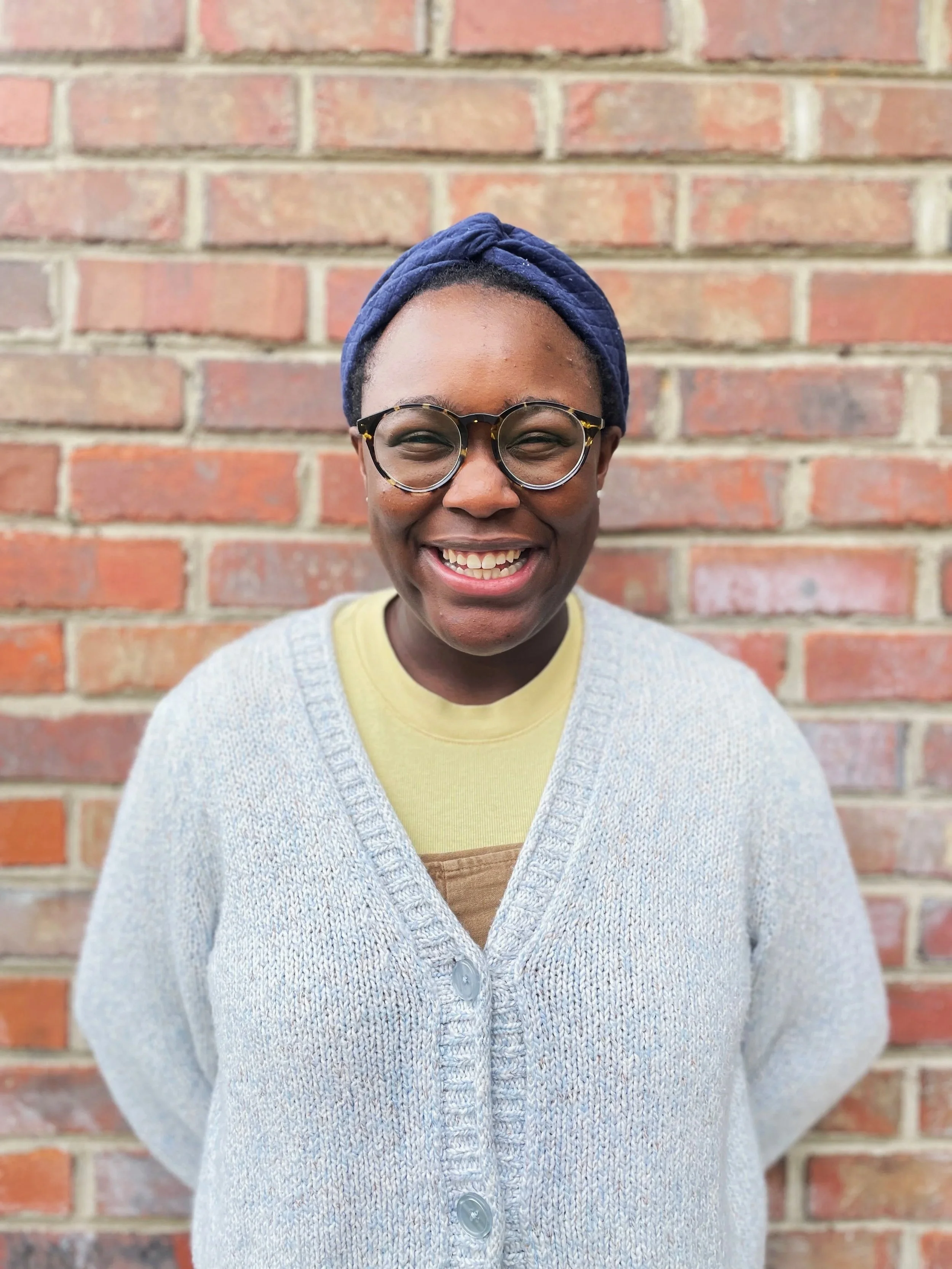 A woman with dark skin wearing glasses, a blue head wrap, and a light gray cardigan, smiling in front of a brick wall.