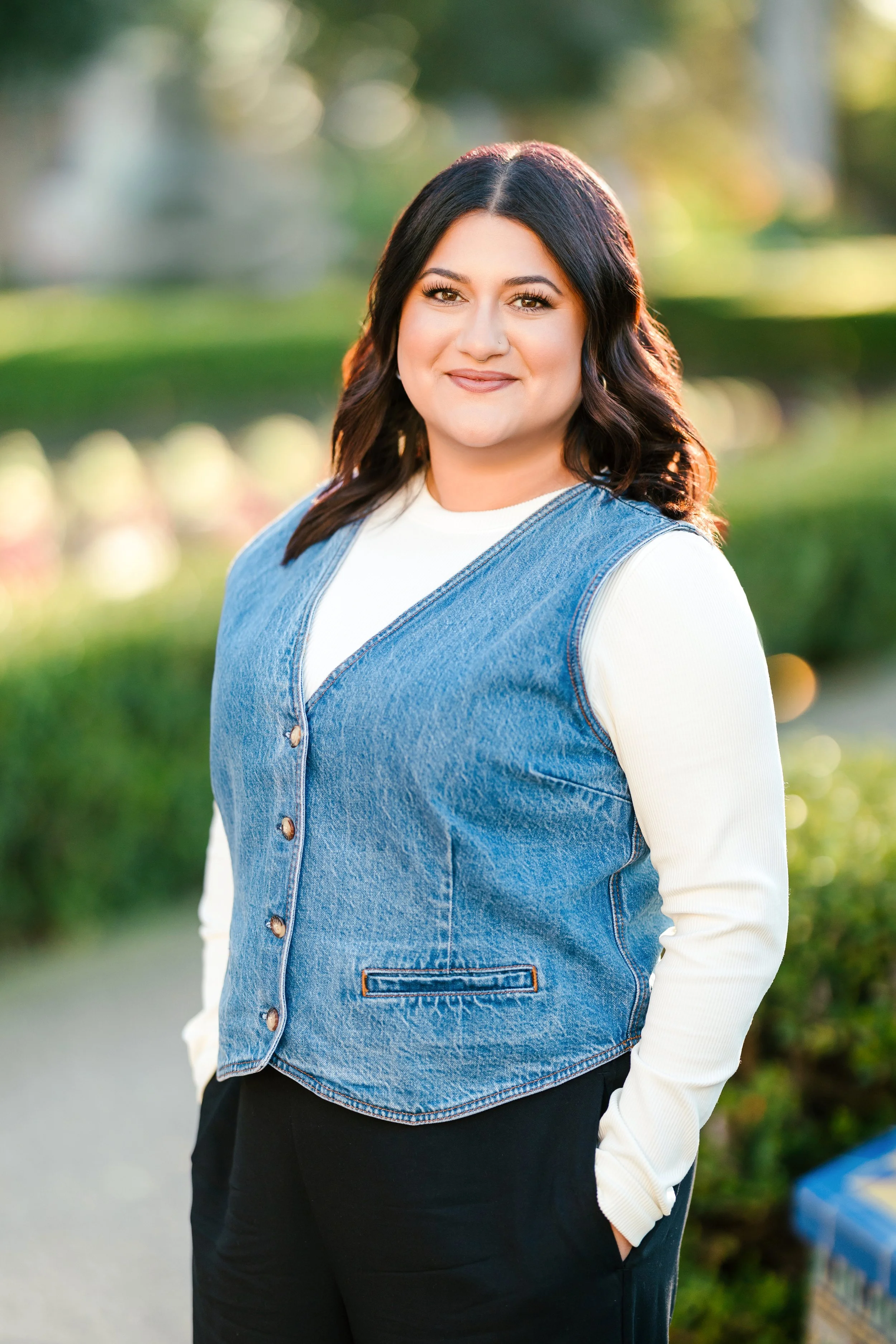 Portrait of a woman standing outdoors, wearing a white long-sleeve shirt and a blue denim vest, with her hands in her pockets. She has shoulder-length dark hair and is smiling. The background is blurred greenery and sunlight.