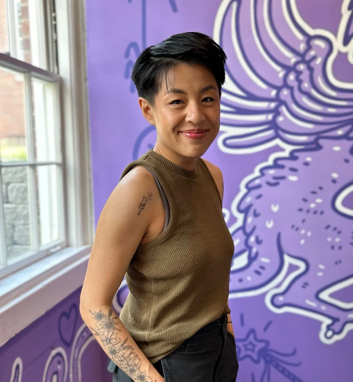 Therapist with short black hair and tattoos on her arm, wearing a sleeveless brown top, smiling and standing in front of a purple mural with a dragon design.