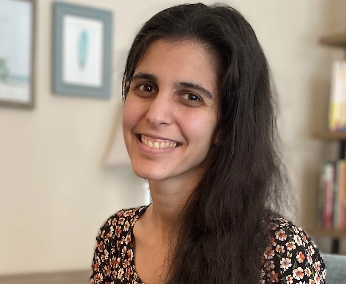 A woman with long dark hair, smiling, wearing a floral top, in a room with framed pictures and a bookshelf in the background.