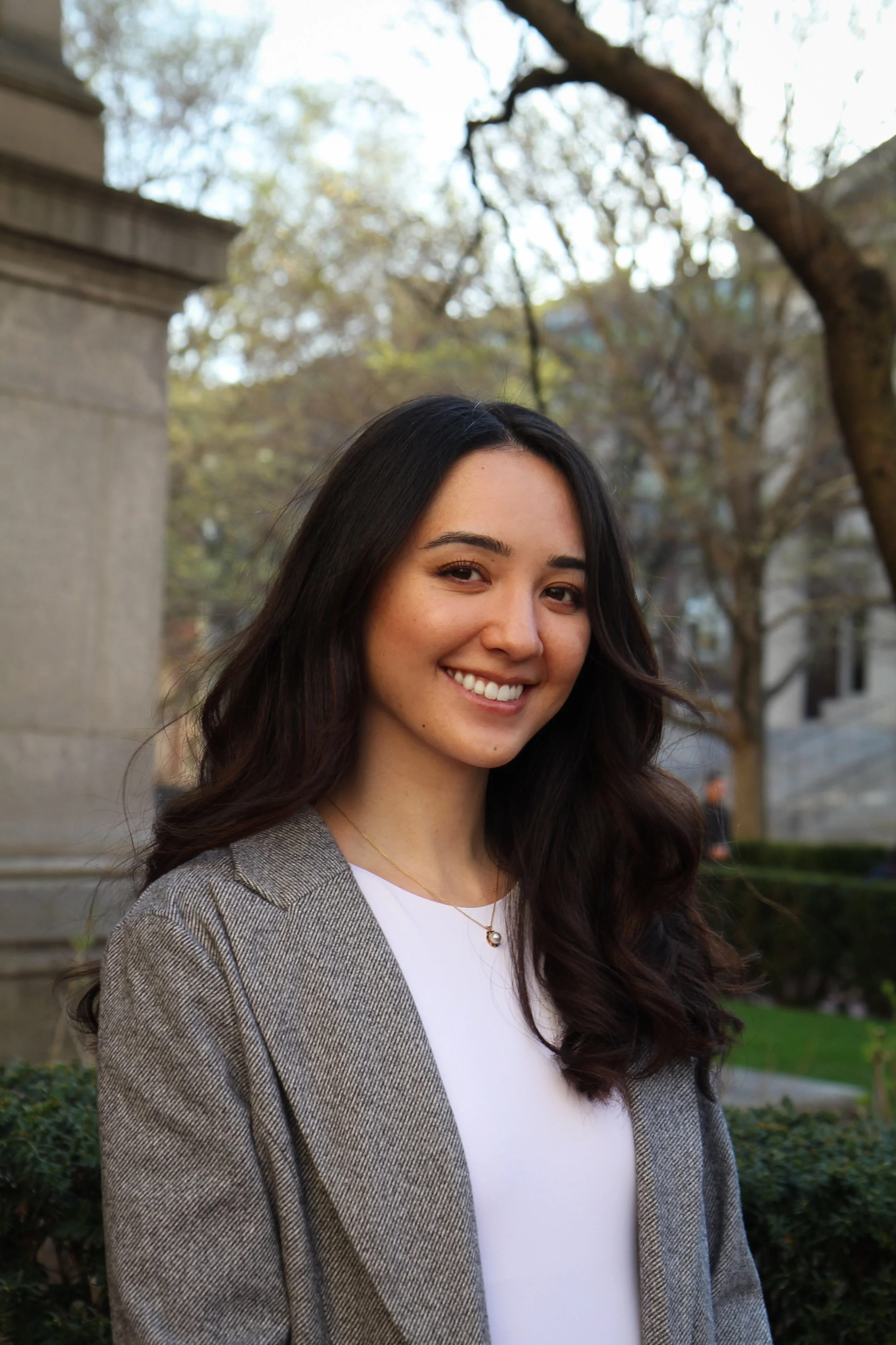 A smiling woman with long dark hair outdoors in front of trees and a stone structure, wearing a gray blazer and a white top.