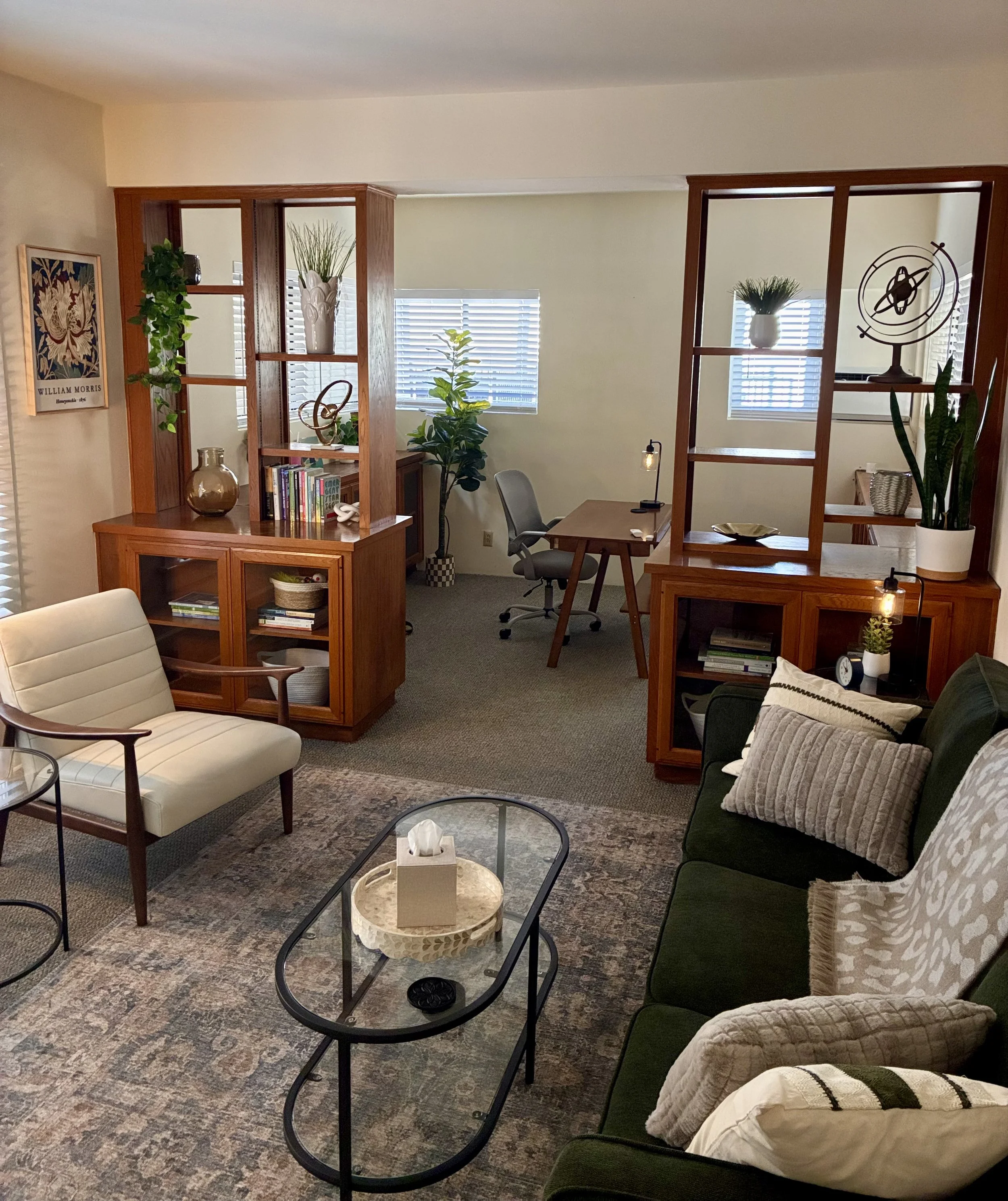 Living room with beige walls, wooden shelves, green and light-colored furniture, featuring plants, books, and decorative items. Natural light streams through window blinds, and a cozy atmosphere is enhanced with patterned pillows and a textured rug.