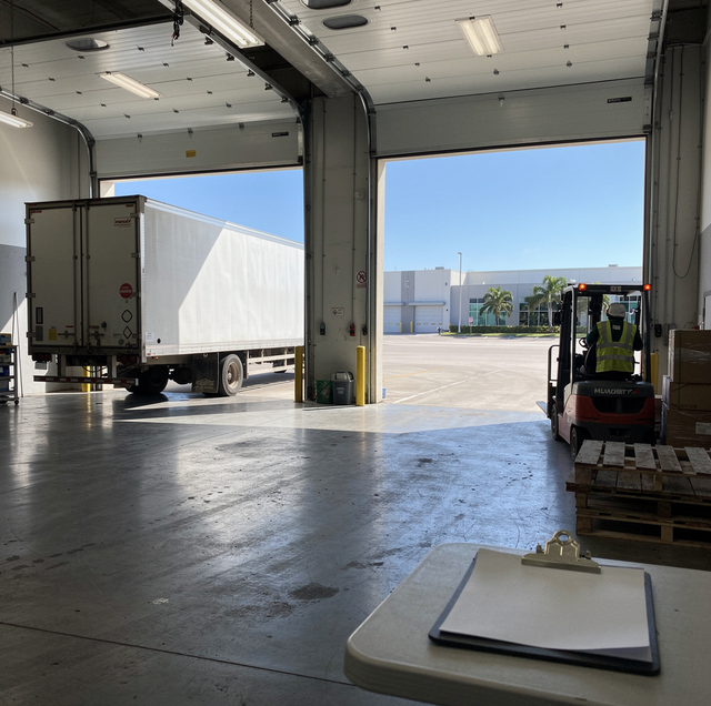 Interior view of an open garage looking out to a concrete lot with palm trees and a white building under blue sky.