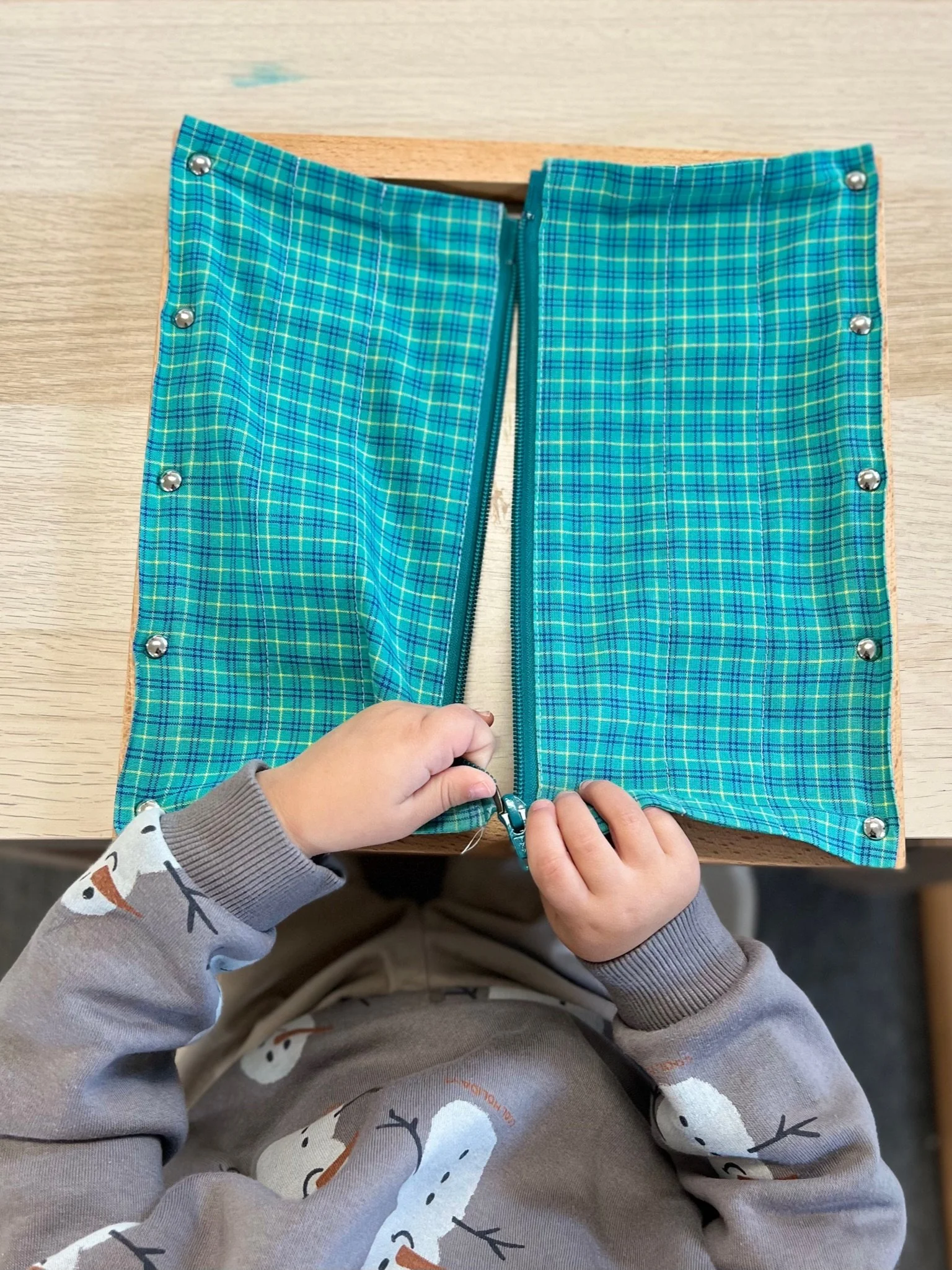 A young child in a snowman-patterned sweater zipping up a plaid fabric cover with metal snaps and a zipper, placed on a wooden table.