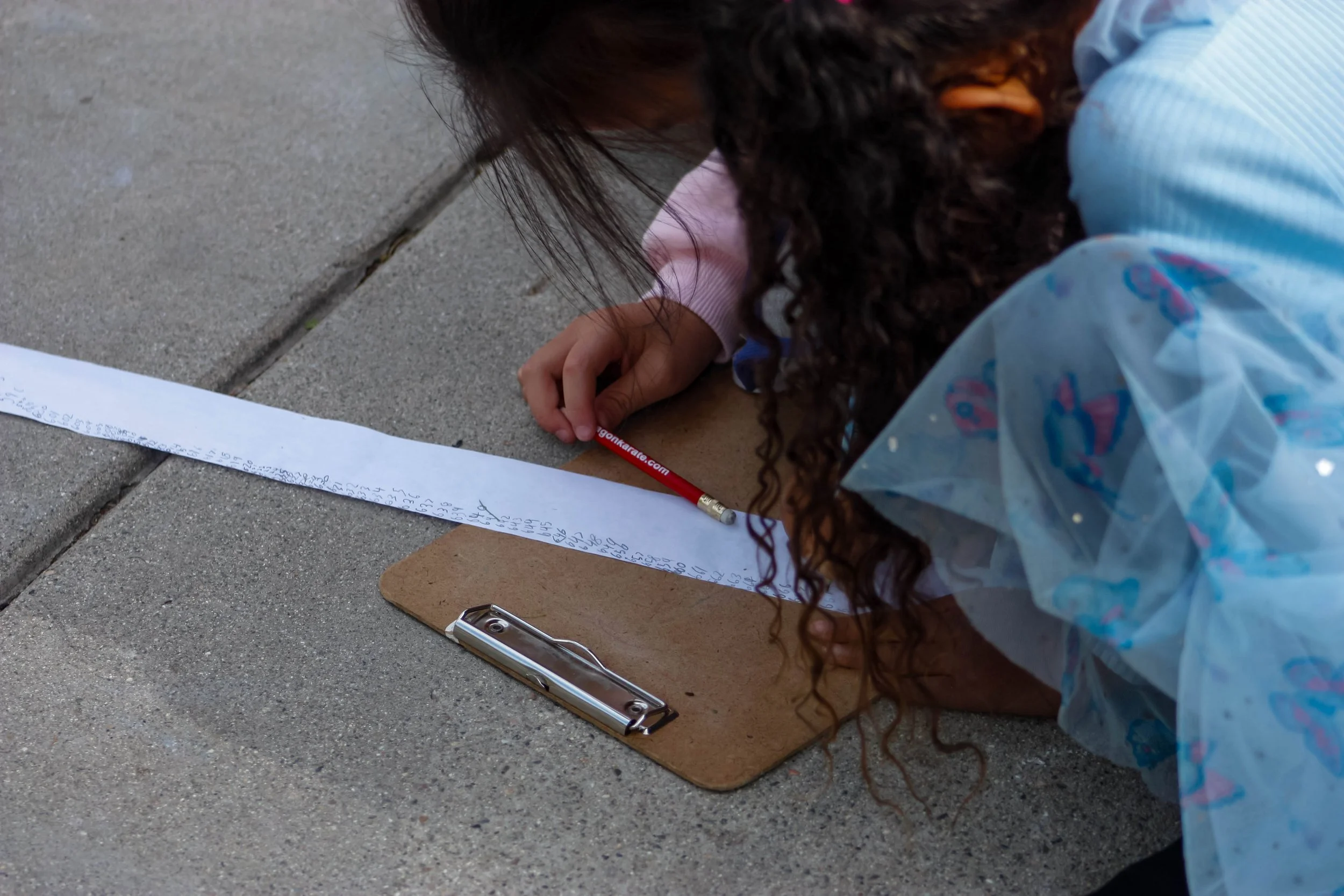 Child writing on a long strip of paper on the sidewalk with a pencil, surrounded by a clipboard and wearing a colorful dress.