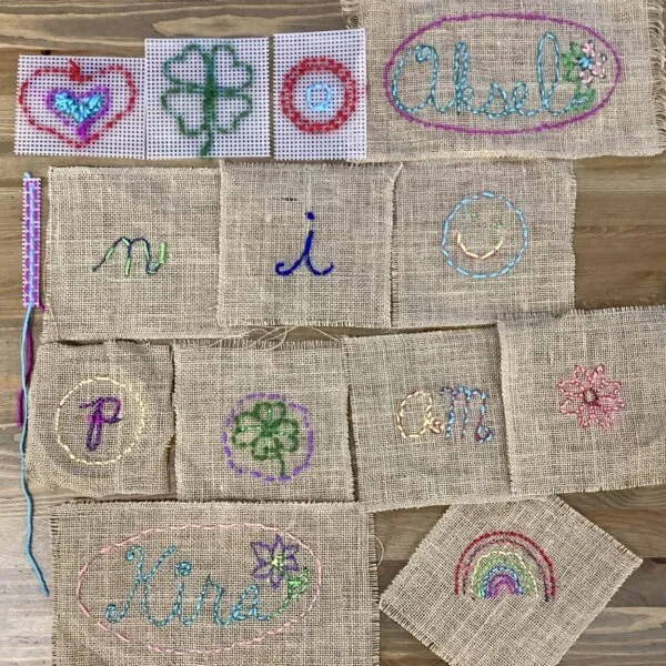 Various pieces of burlap fabric featuring colorful embroidered words and designs, including hearts, rainbows, and floral patterns, arranged on a wooden surface.
