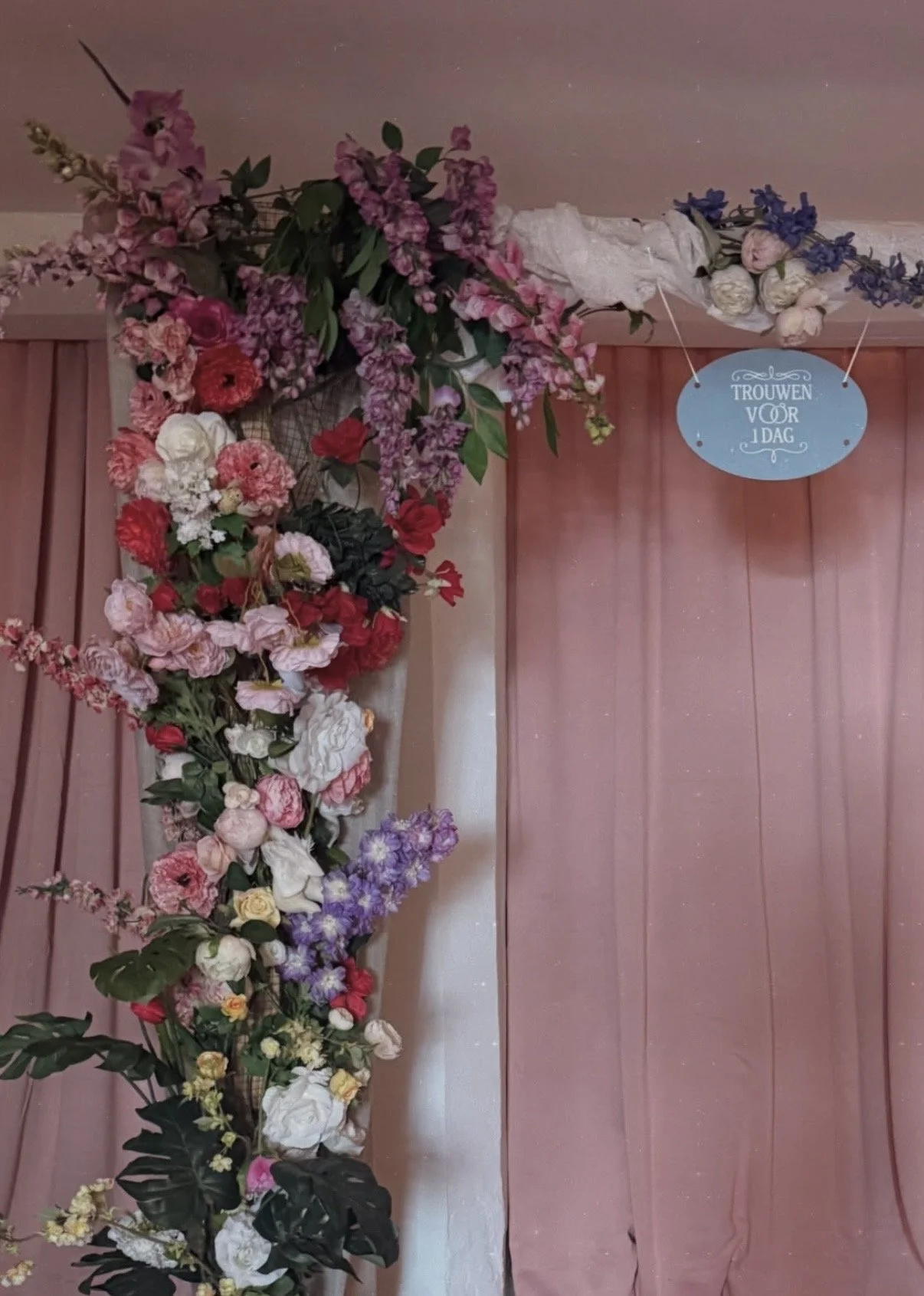 Bloemenkrans met verschillende roze, witte, rode, paarse en blauwe bloemen, hangend aan een plafond, met een lichtblauw bordje met witte tekst 'Trouwen voor 1 dag' eraan bevestigd.
