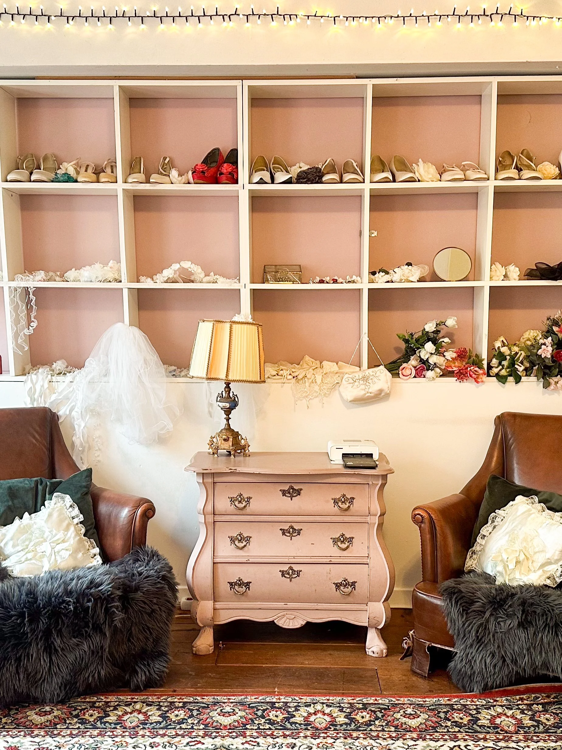 Interieur met houten vloer, twee leren fauteuils met kussens en bonten voeten, een roze antieke ladekast met een lamp en papier op bovenkant, een witte lockersysteem met schoenen en accessoires, een vloerkleed, en muurdecoratie voor een bruiloft zoals bloemen, een spiegel en een trouwhoed.