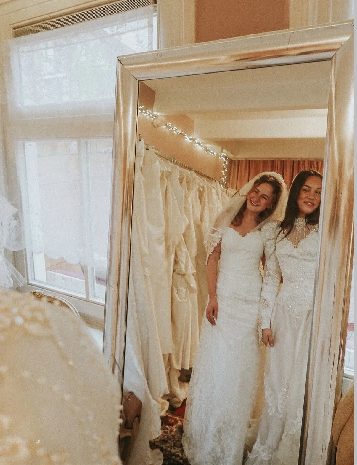 Twee vrouwen in witte trouwjurken staan voor een grote spiegel, waarin ze elkaar glimlachend kijken, op een kamer met gekleurde muren en decoraties, omringd door trouwjurken.