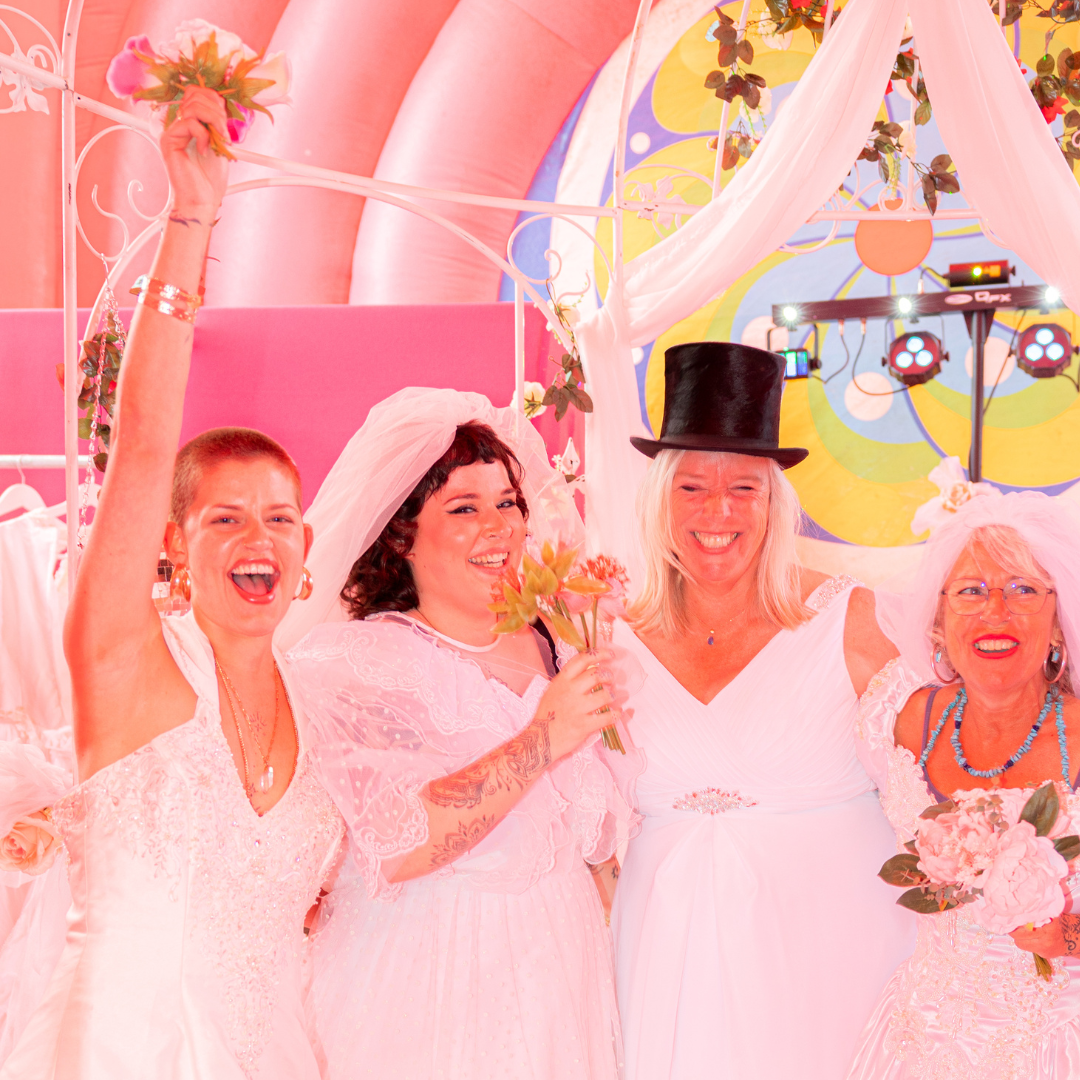 Vier vrouwen op een feest, gekleed in witte jurken, met bloemen en accessoires, lachend en feestend onder kleurrijk decor.