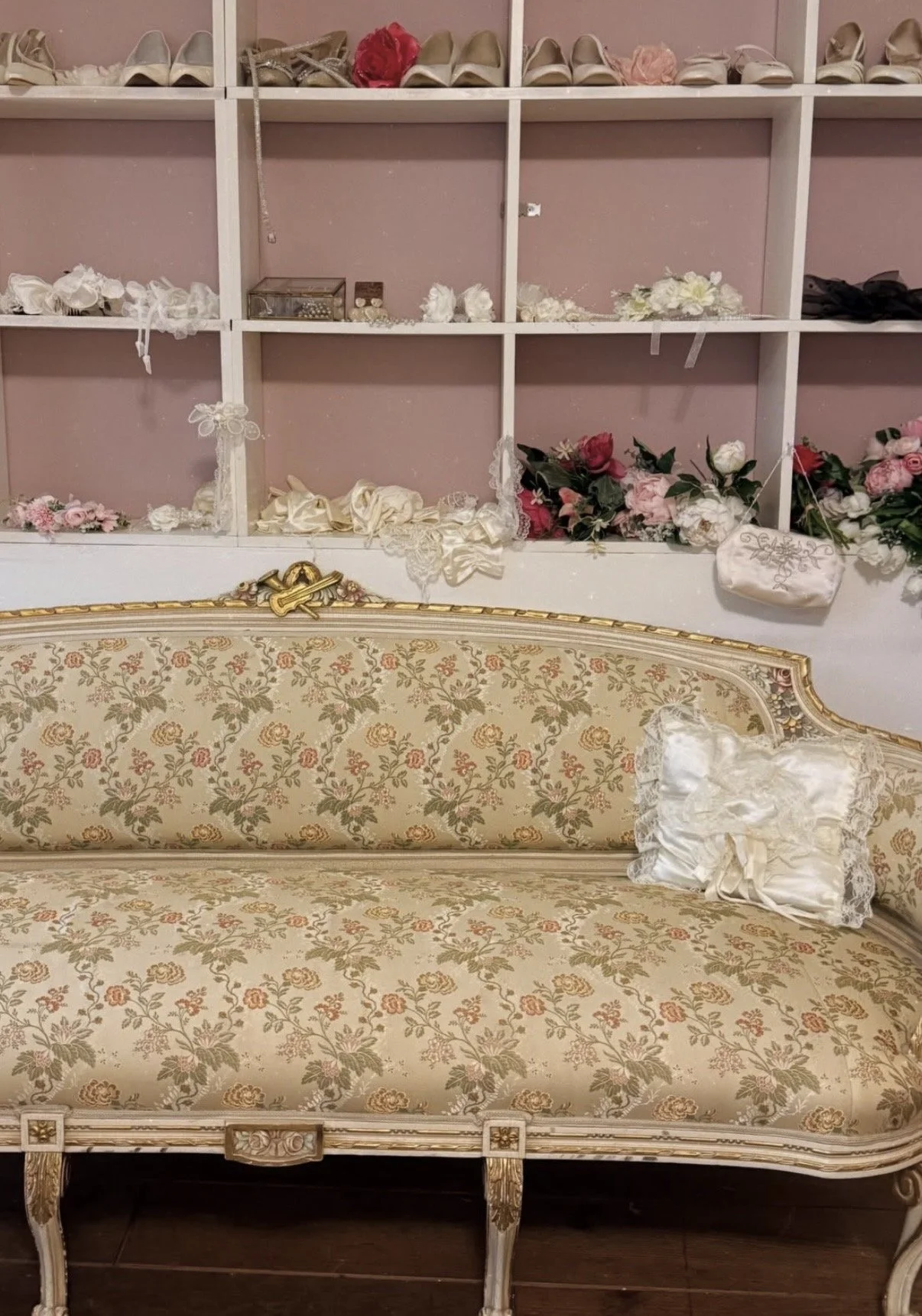 Een antieke bank met gebloemde bekleding en gouden details, met een sierkussen op de armleuning, voor een muur met opbergkubussen gevuld met schoenen, bloemen en accessoires.