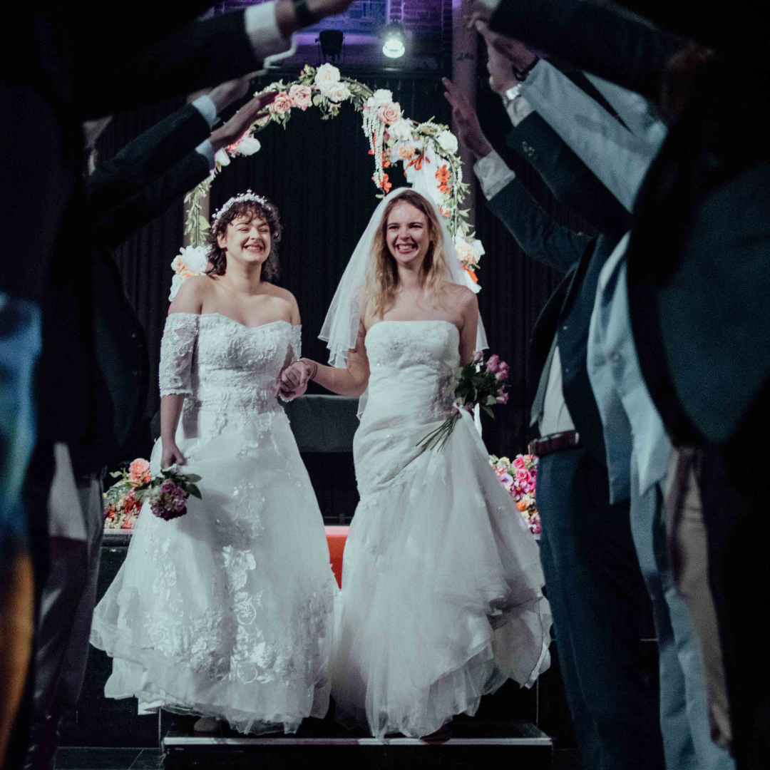 Twee bruidspaartjes in trouwjurk houden handen vast, omringd door gasten die met hun armen een tunnel vormen tijdens de bruiloftceremonie. De bruiden lachen en staan onder een bloemenboog. De achtergrond is donker met bloemendecoratie.