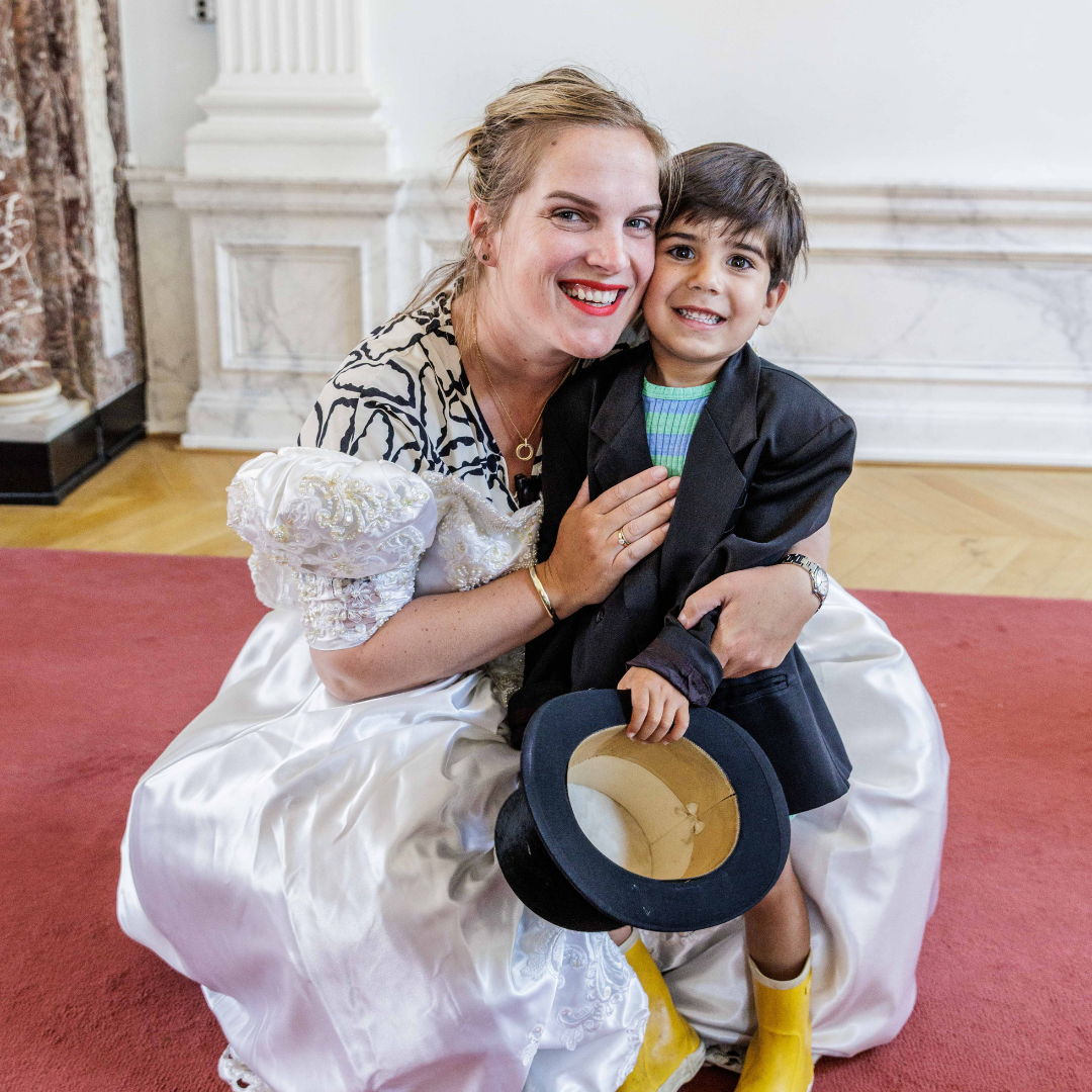 Een vrouw en een jongen knuffelen samen in een stijlvolle kamer, beide glimlachend. De vrouw draagt een witte jurk met bloemkraag en de jongen draagt een regenjas en gele laarzen, terwijl hij een hoed vasthoudt.