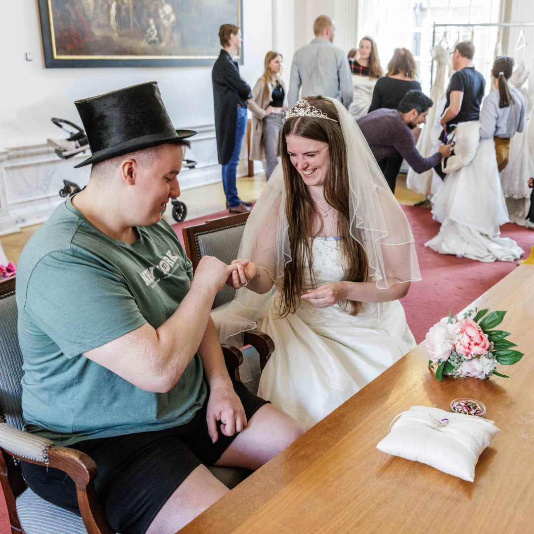 Gelukkig bruid en bruidegom op een bruiloft, de bruidegom met een hoed en de bruid met een tiara en sluier, ze lachen en hand in hand op een houten tafel met bloemen en sieraden, meerdere mensen in de achtergrond in een lichte ruimte met grote ramen.