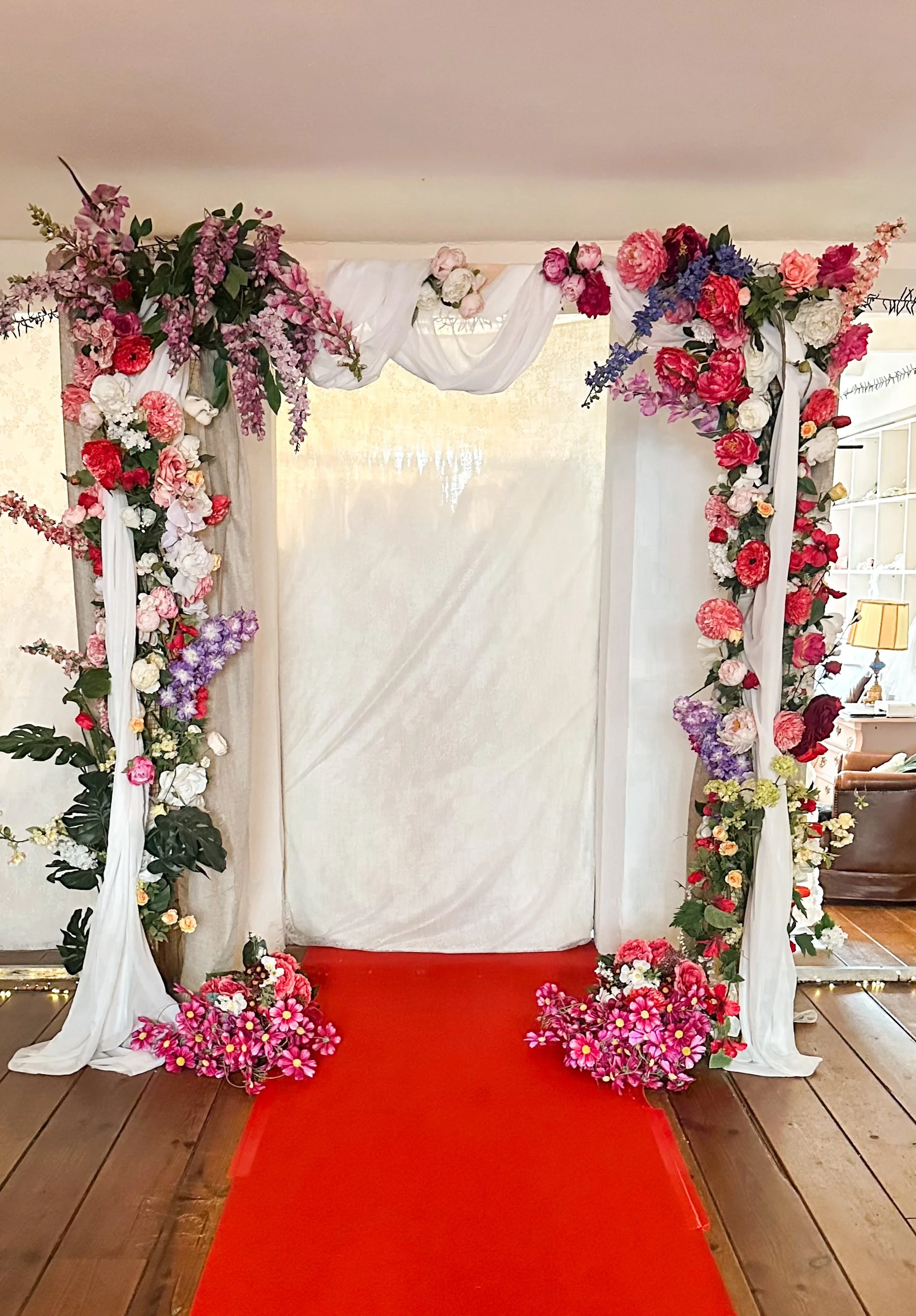 Een bloemeningang met een rode loper en een bloemenboog, versierd met roze, paarse en witte bloemen en witte doeken.