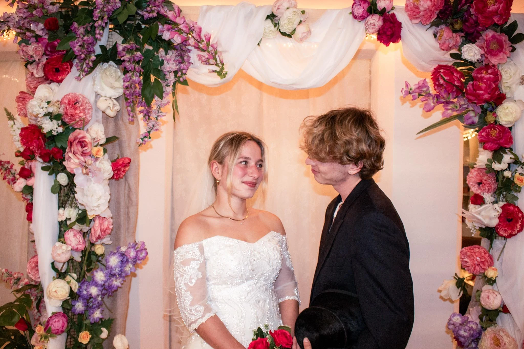 Een paar dat net gaat trouwen staat voor een bloemenboog met roze, witte en paarse bloemen tijdens een huwelijksceremonie.