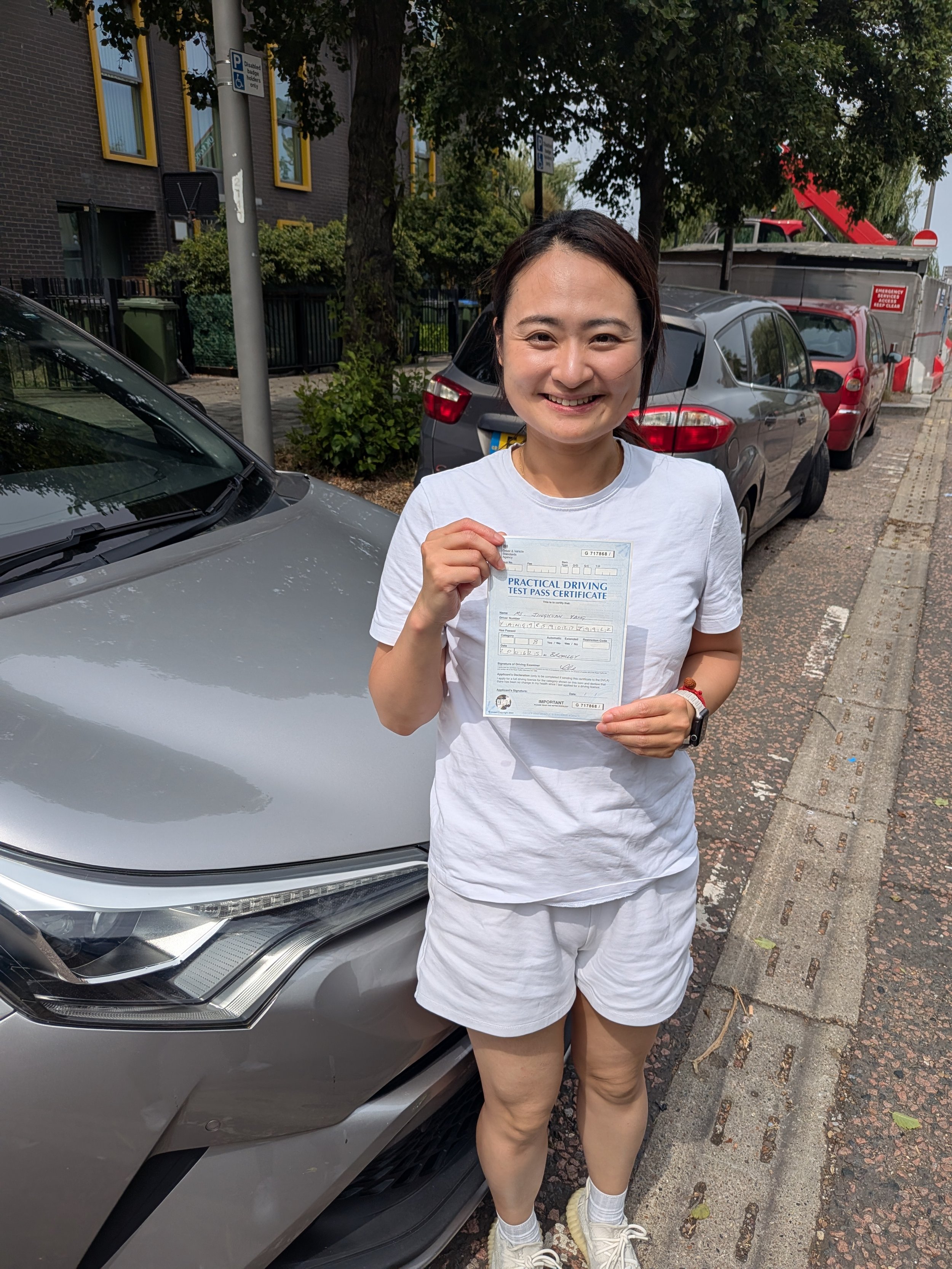 Woman standing on city street holding a practical driving test pass certificate, smiling, with parked cars and trees in background.