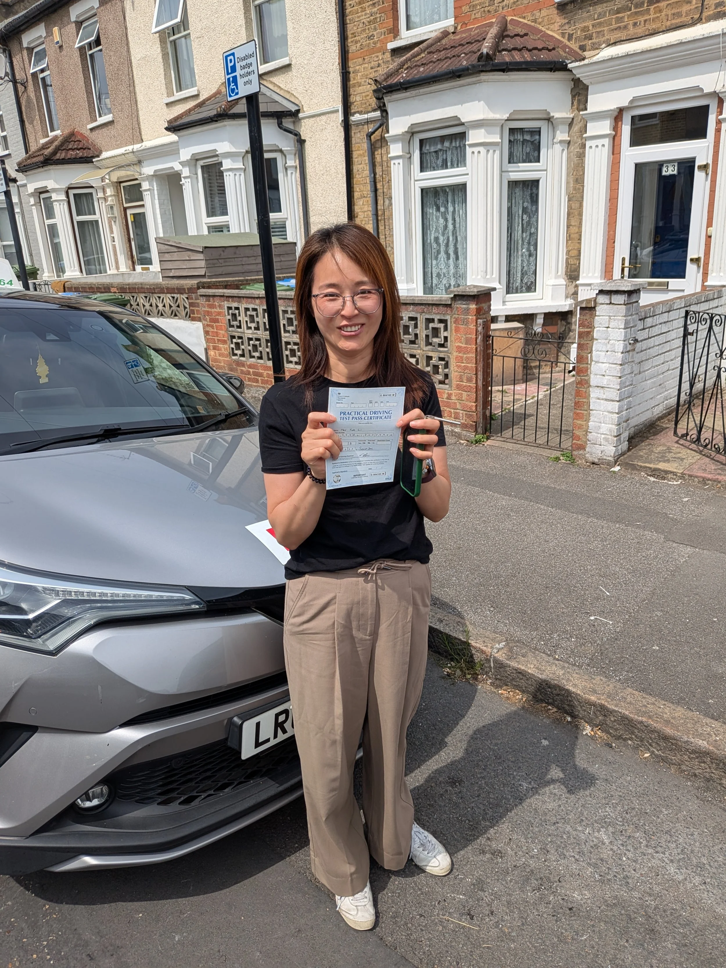A woman standing next to a silver car on a residential street, holding a driving test certificate and smiling. She is wearing glasses, a black t-shirt, beige pants, and white sneakers.