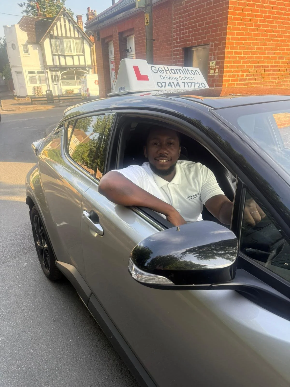 A man smiling while sitting in the driver's seat of a gray driving school car. The car has a sign on top that reads 'GoHamilton Driving School' with a phone number. The man is wearing a white polo shirt with the driving school's logo.