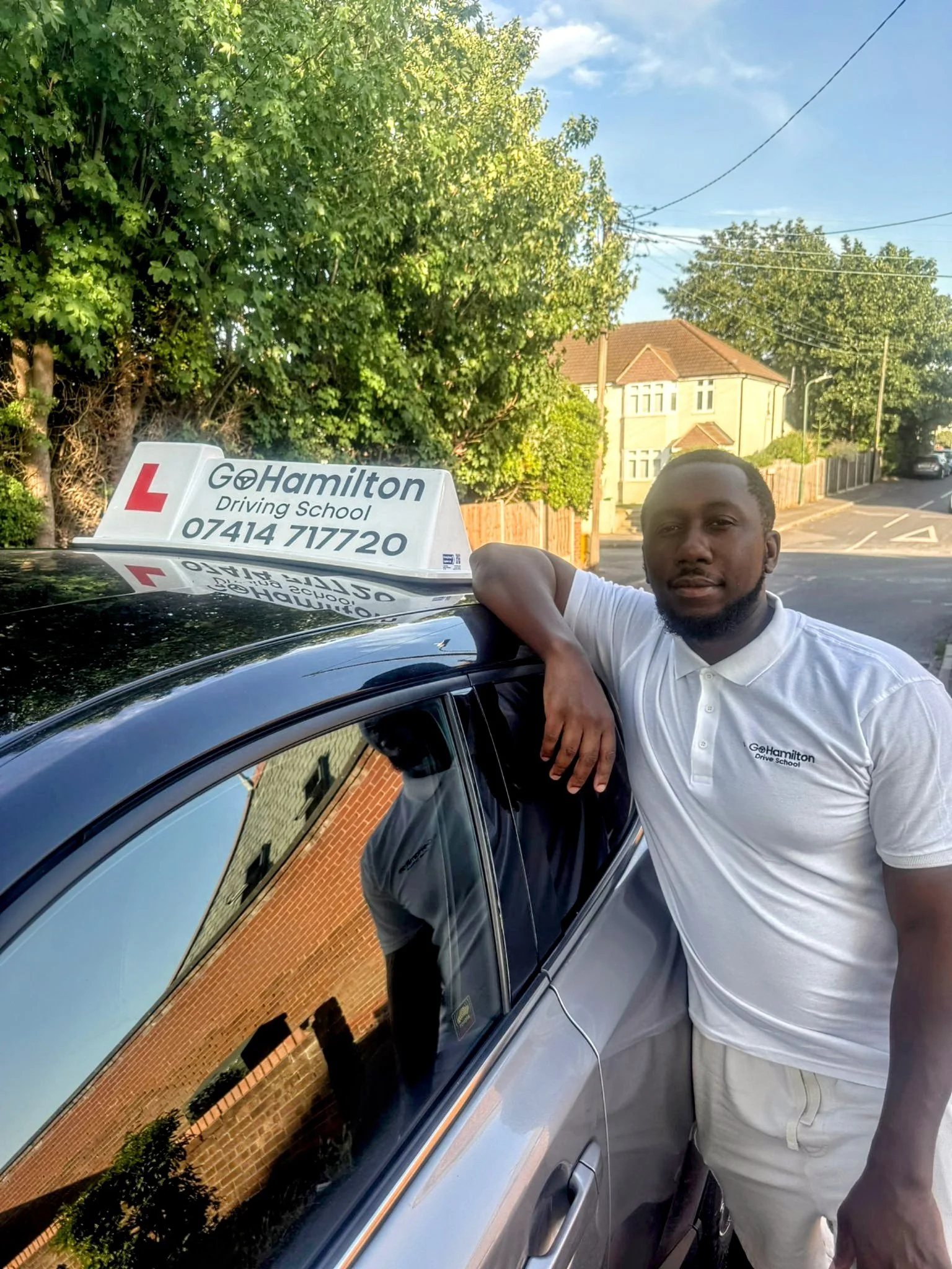 A man stands leaning on a black car with a driving school sign on top that reads 'G Hamilton Driving School' with a phone number. The man is wearing a white polo shirt and beige pants, and is outdoors with trees, houses, and a street in the background.