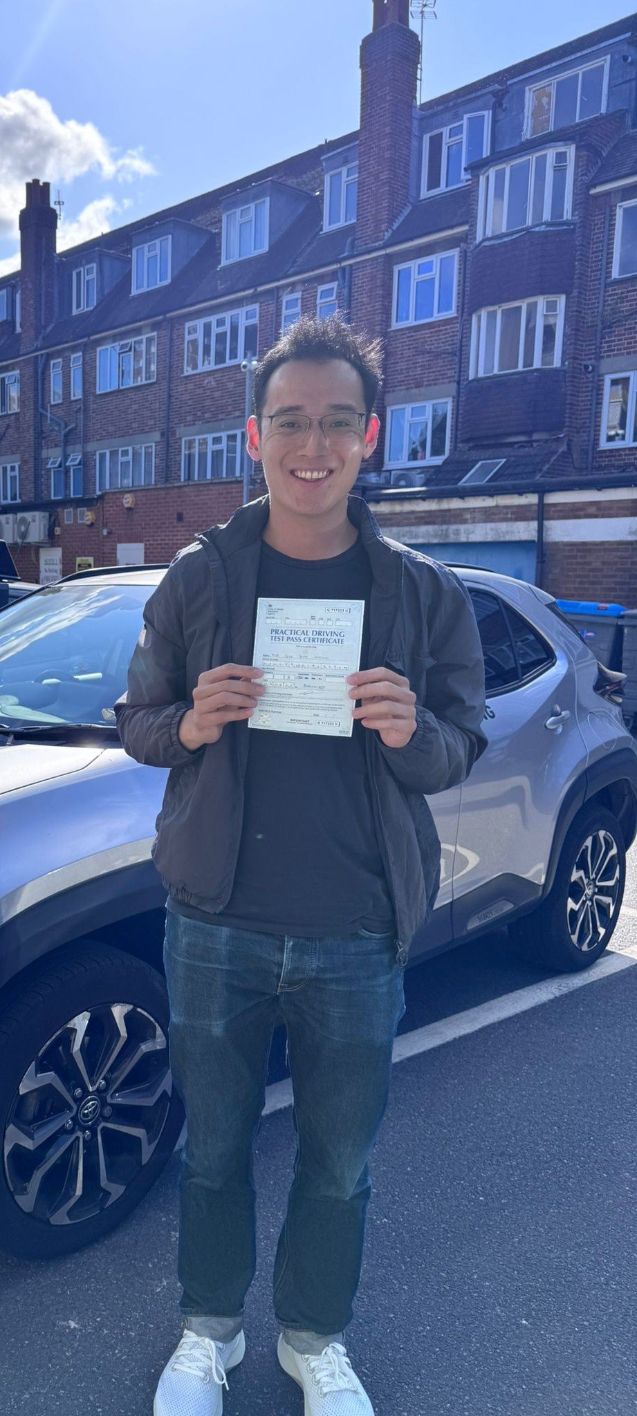 A young man holding a practical driving test pass certificate, standing outdoors in front of a silver car on a city street, smiling.